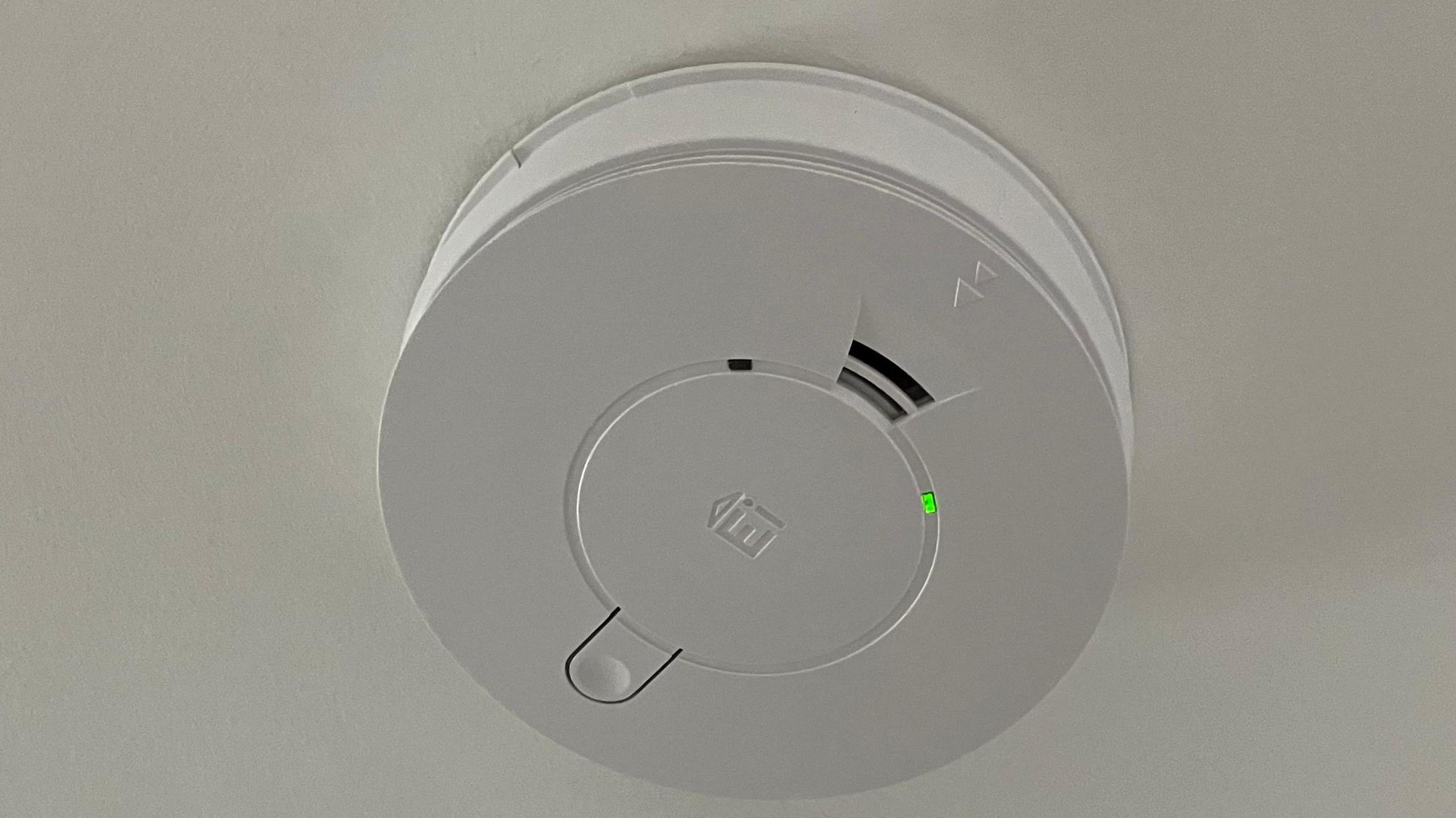A plastic circular carbon monoxide detector on a white painted ceiling. There is some vents in it and a button. There is also a small green light.
