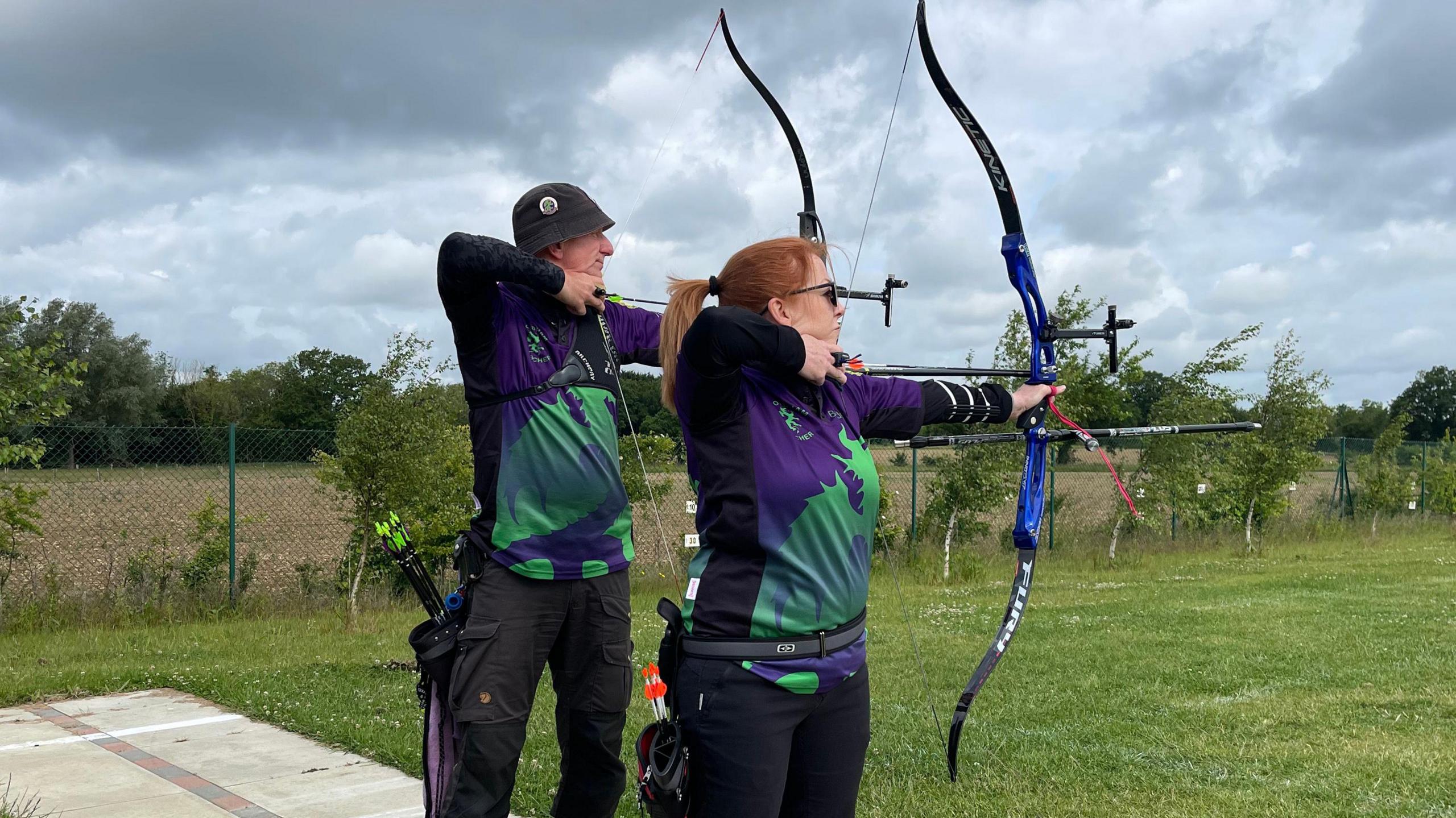 Two people on the shooting line at an archery club with bows and arrows who are both stood sideways ready to hit a target.