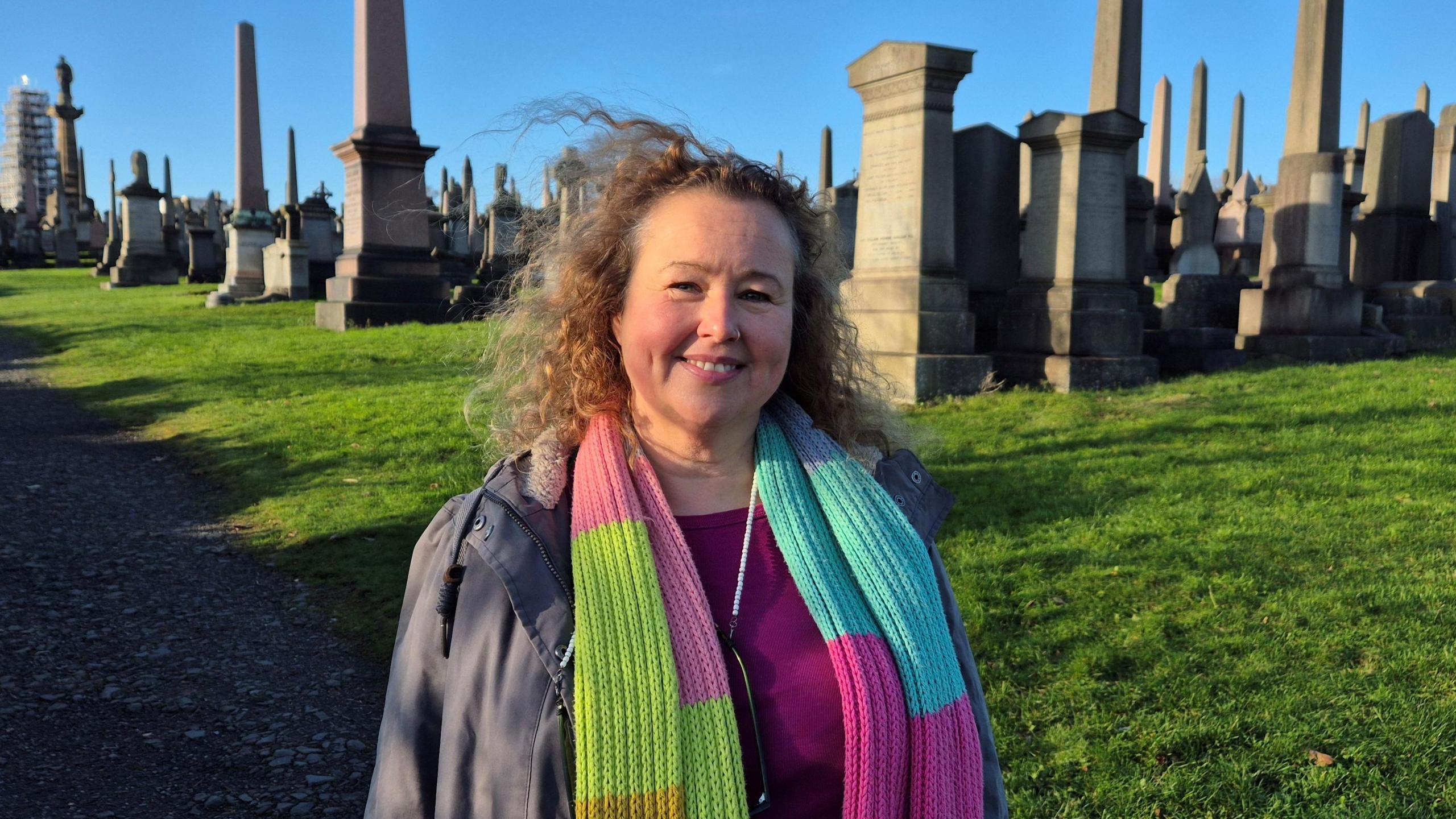 Onya Attridge, who has light hair, is wearing a jacket and multi-coloured scarf  in the graveyard on a sunny day.