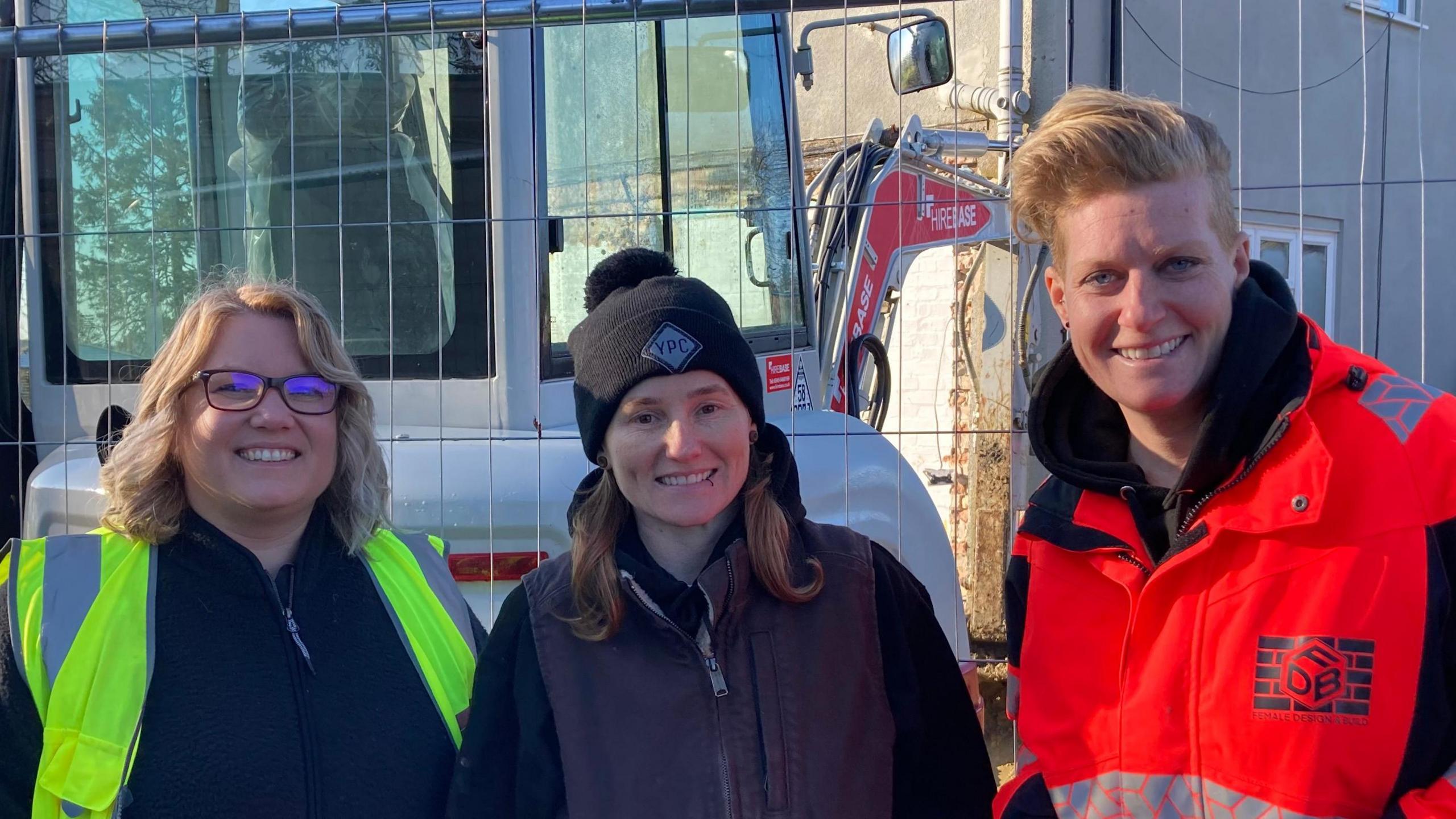 Marta Holton, Yas Poole and Kat Parsons smile at the camera in front of a construction site. Marta wears a yellow hi-vis jacket and see has shoulder length blonde hair. Yas wears a black beanie hat and black coat while Kat has short blonde hair and wears an orange hi-vis coat. 