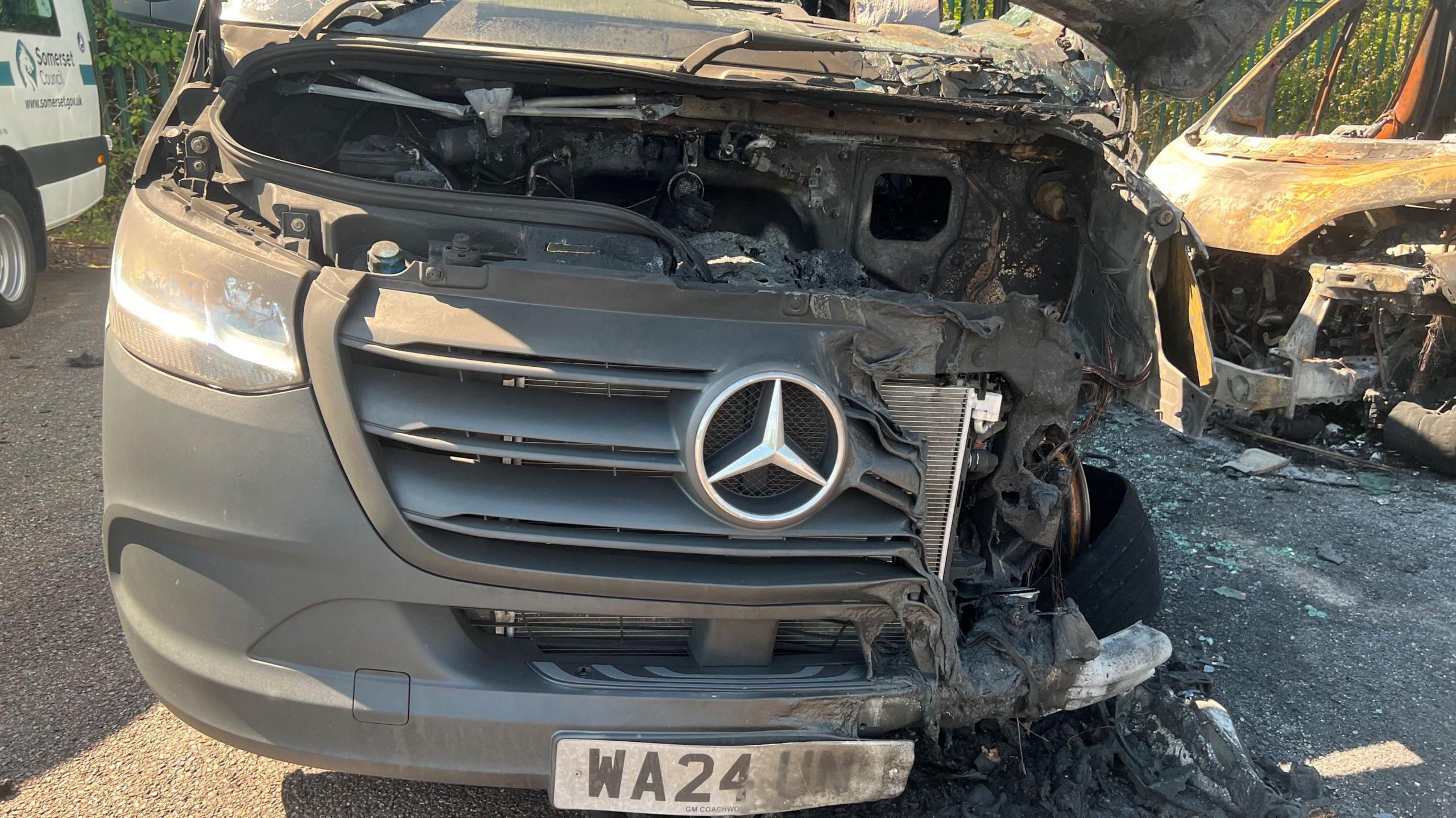A black Mercedes minibus pictured up close from the front. The left hand side of the front has been burnt and melted away.