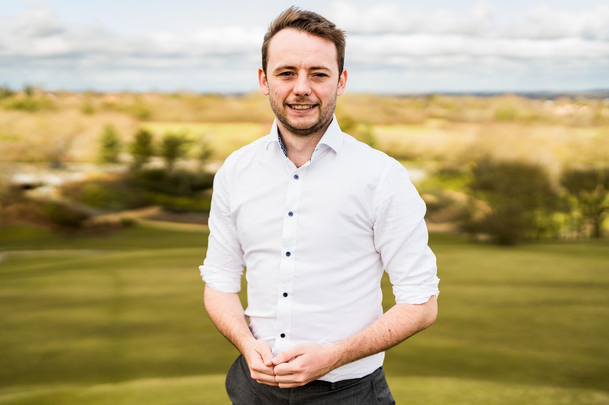 Labour MP for MK North, Chris Curtis, standing in green space outside in a white shirt. He has short brown hair and stubble and is smiling at the camera.