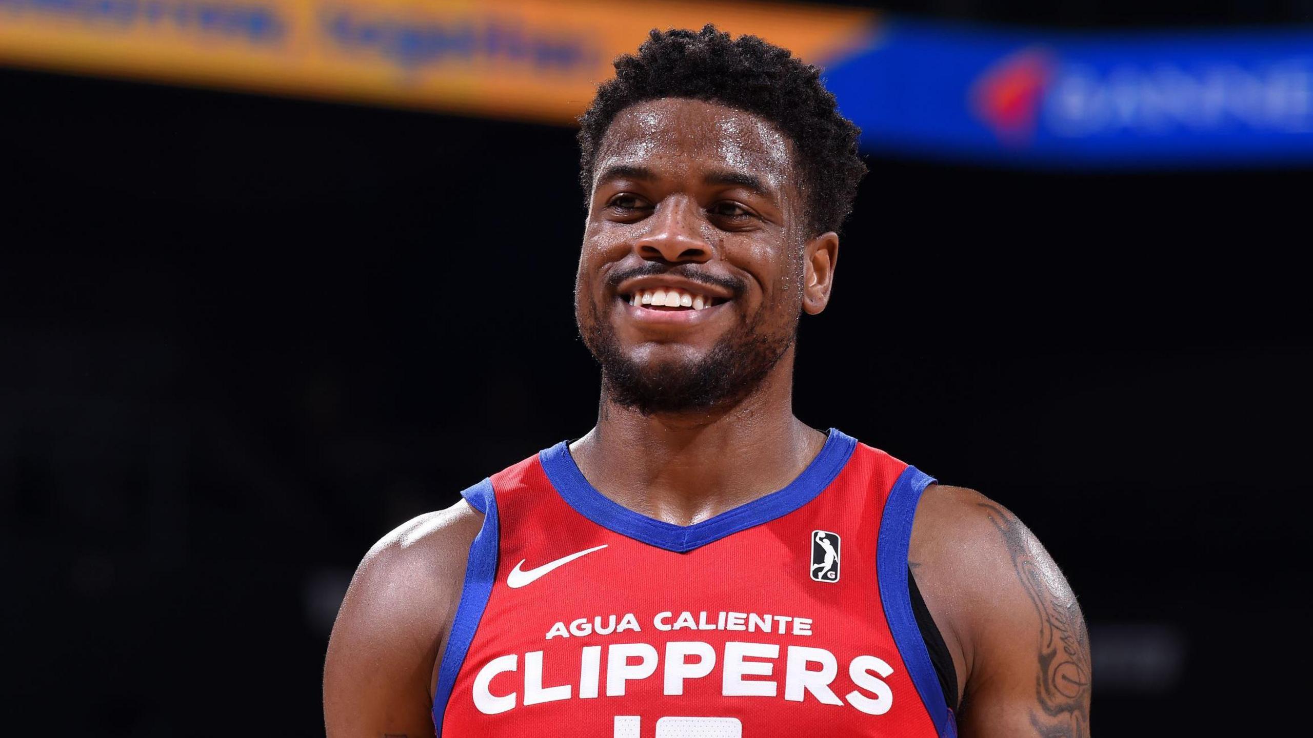 Malik Dunbar smiles while on court in a basketball game. He is wearing a red sports kit with Agua Caliente Clippers written on it in white typeface.