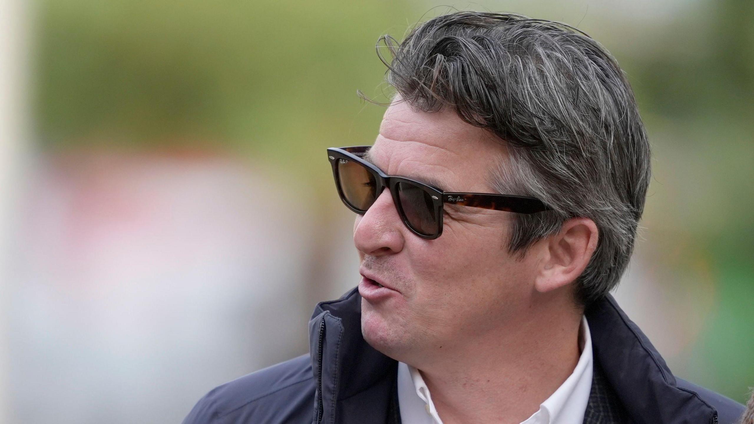 Joey Barton smiles as he speaks to someone over his shoulder on his way into court. He has shirt dark hair and wears dark shades.