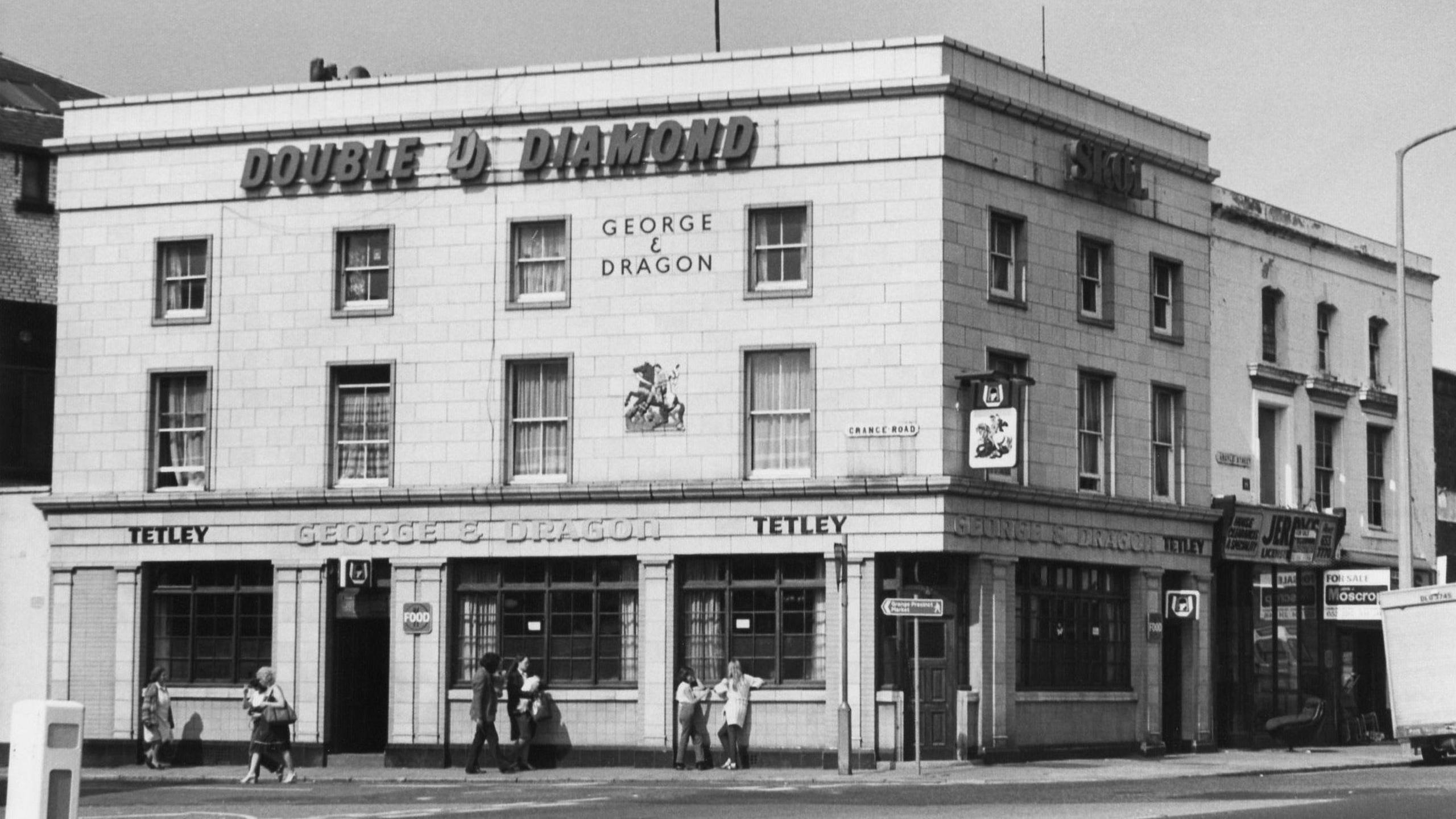 Black and white image of The George and Dragon pub, which is a three-storey white building on a junction. Some people can be seen standing or walking past it.