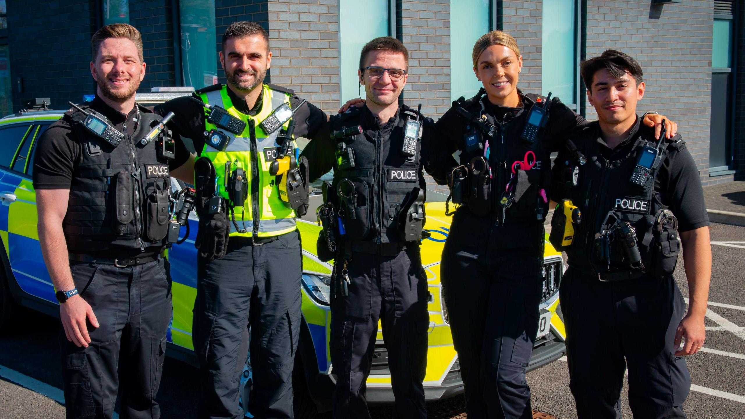 Five Avon and Somerset Police officers - four male and one female - stand with their arms linked facing the camera. Three of them are in dark uniforms with radios and other kit attached to their jackets, while one has a high-vis vest on. Behind them is a police car with a blue and yellow livery.