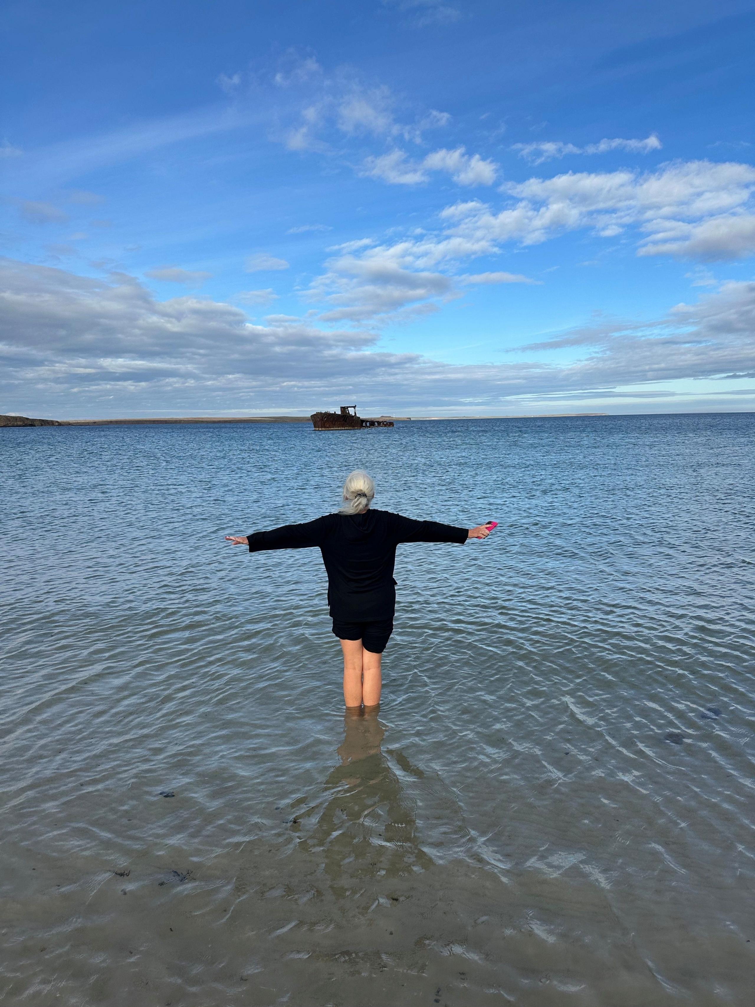 Woman standing in water with her back to the camera and arms outstretched, looking out over the water with a rusted shipwreck in the far distance