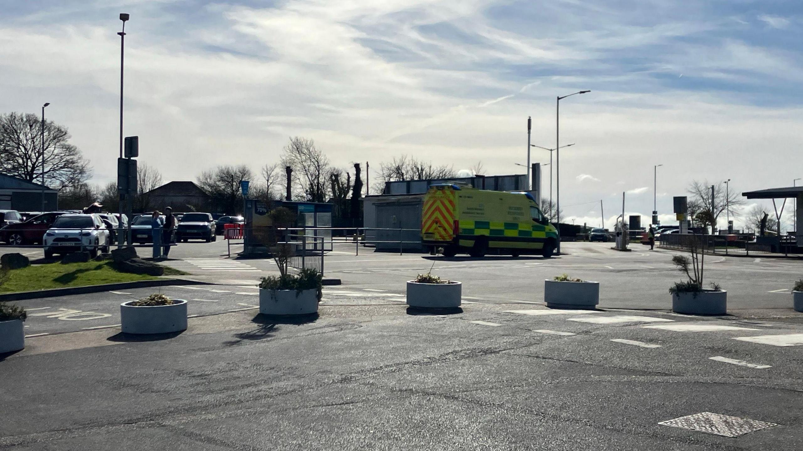 An ambulance parked outside Exeter Airport. Several cars are in a car park next to it. A couple of people are stood near one of the cars. A row of large plant pots are dotted along the road.