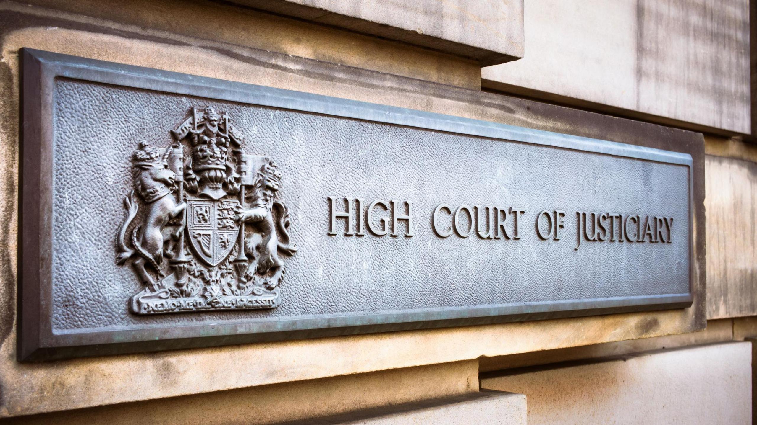 A grey metal sign on an exterior sandstone block wall of the High Court in Edinburgh. The sign has a royal crest and the words "High Court of Justiciary".