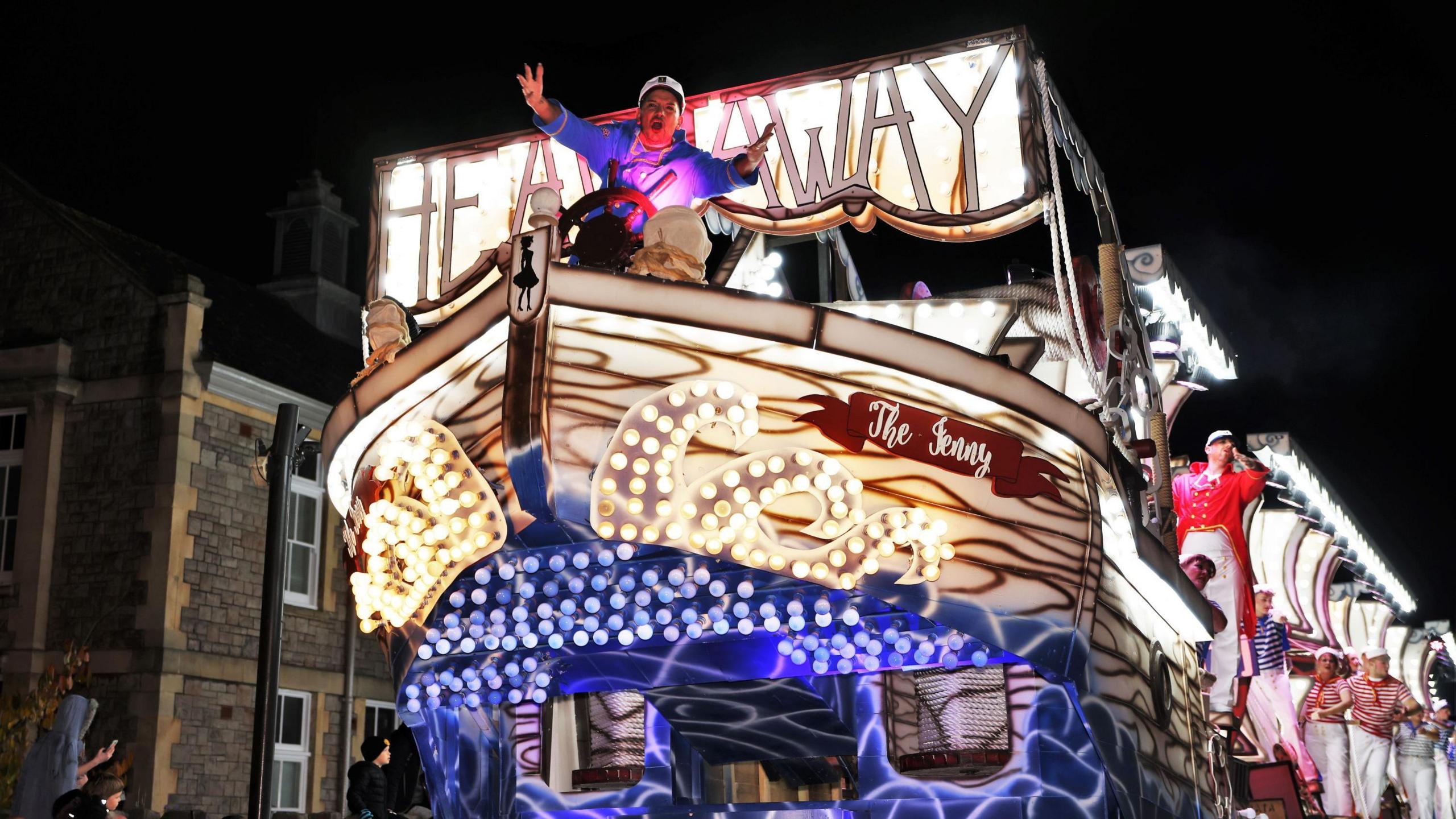 A brightly-lit white, brown and blue carnival cart seen from the front. It is designed to look like a ship and has a captain at the front reaching his arms out towards the camera.