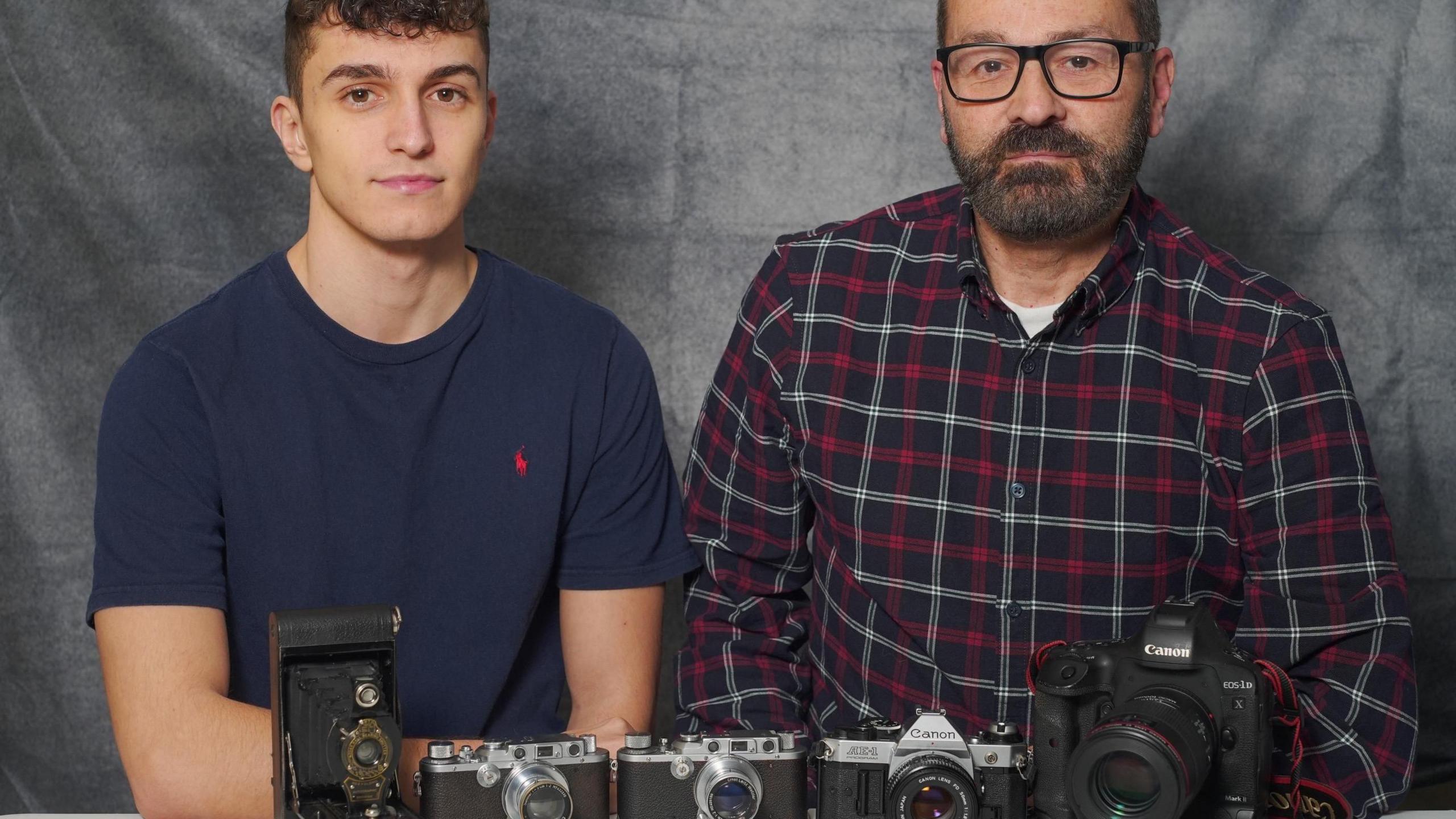 Two men sat a table with a row of cameras, both are looking right at the camera