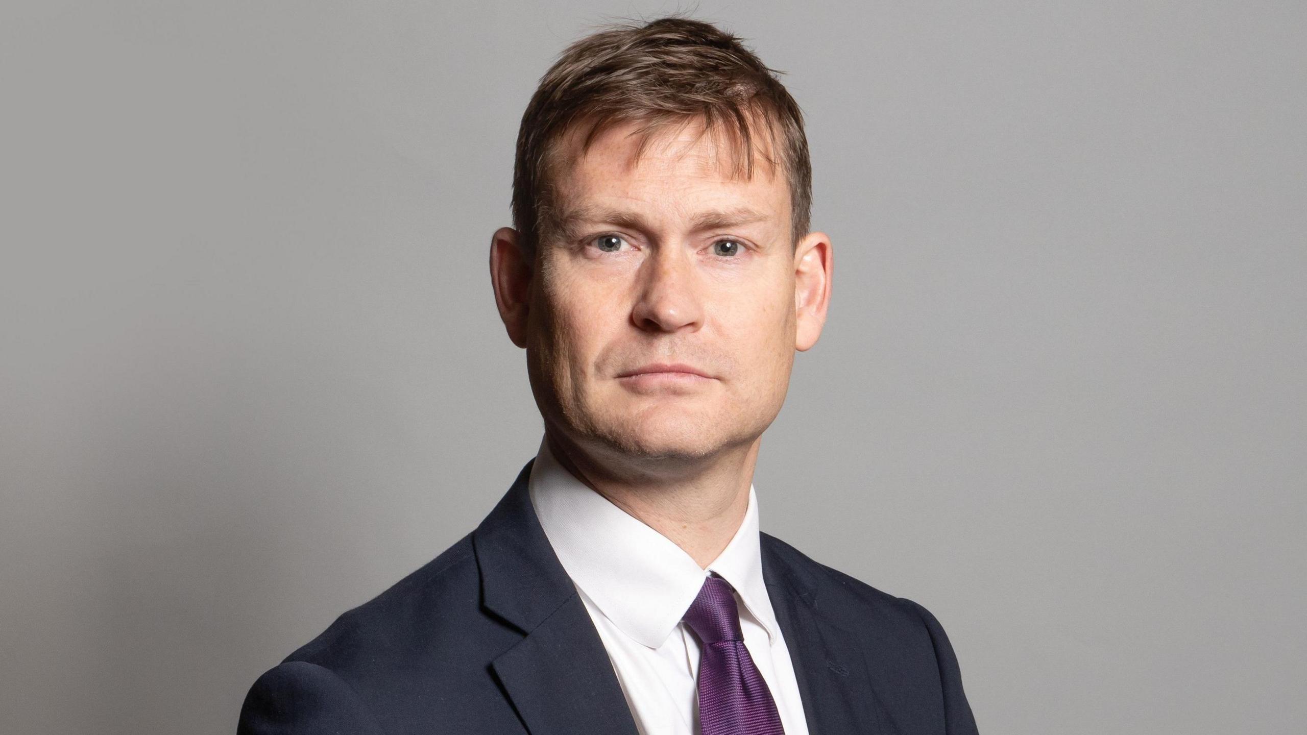 MP Justin Madders posing for an official parliamentary photo, wearing a navy suit and purple tie