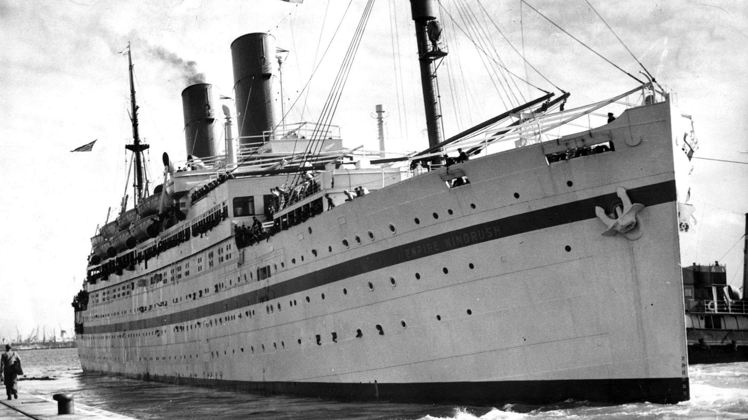 A black and white photograph of HMT Empire Windrush, docked at Southampton in 1954. The ship has a long dark stripe down one side, an anchor at the front and smoke coming from its funnels.