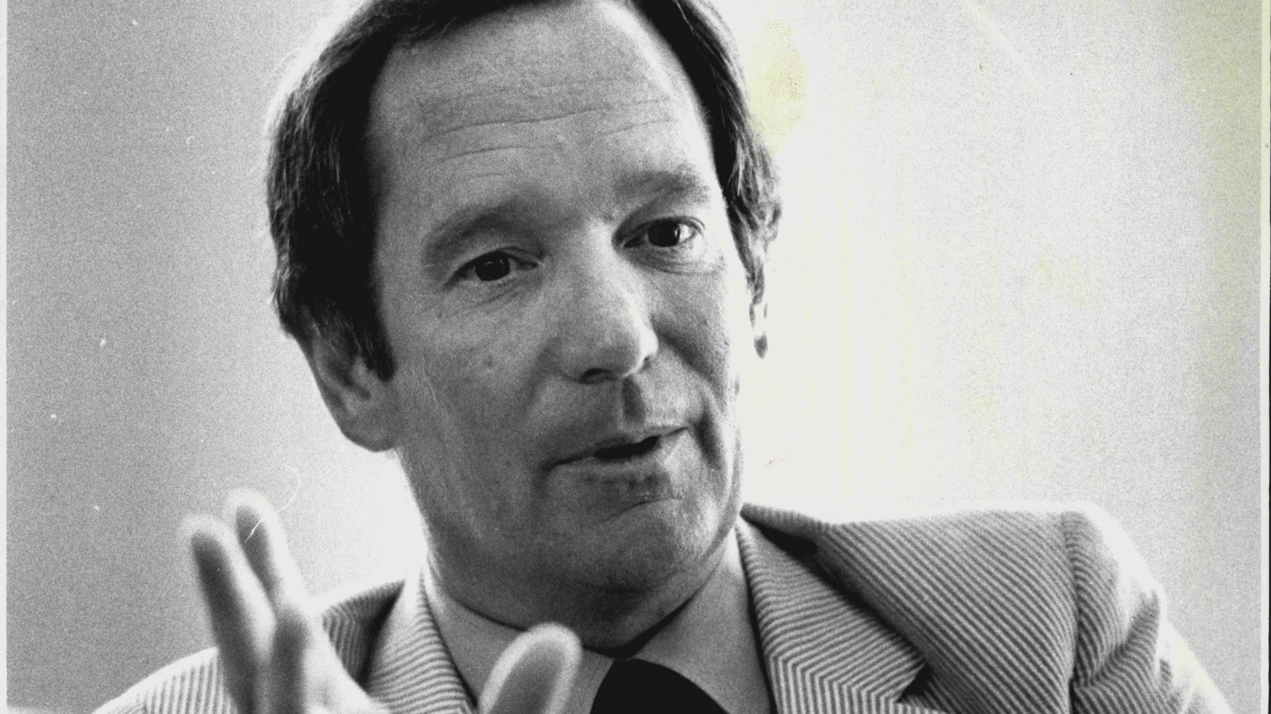 Close up black and white image of broadcaster Michael Buerk. He looks like he is speaking and is gesturing with his right hand. He is wearing a striped suit jacket with a shirt and tie.