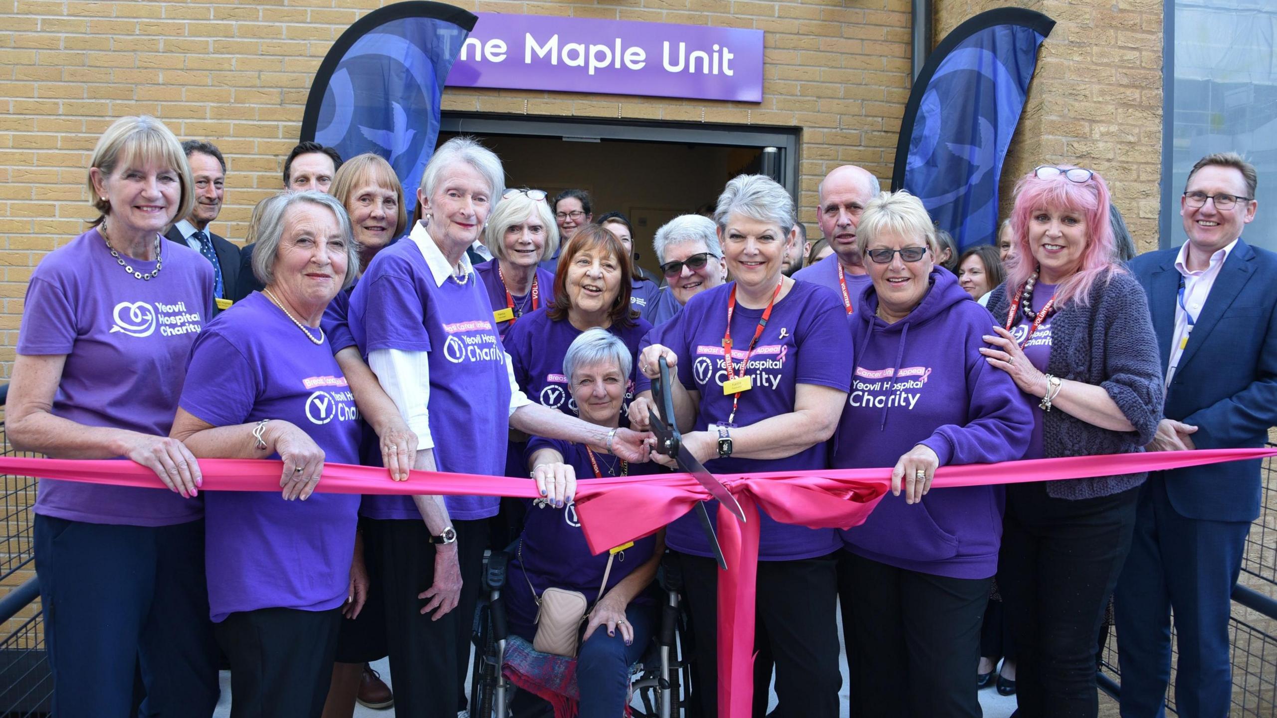 Staff and charity workers at NHS Foundation Trust are standing in front of the new Maple Unit at Yeovil Hospital. They are all wearing purple t-shirts. One of them is cutting a pink ribbon.