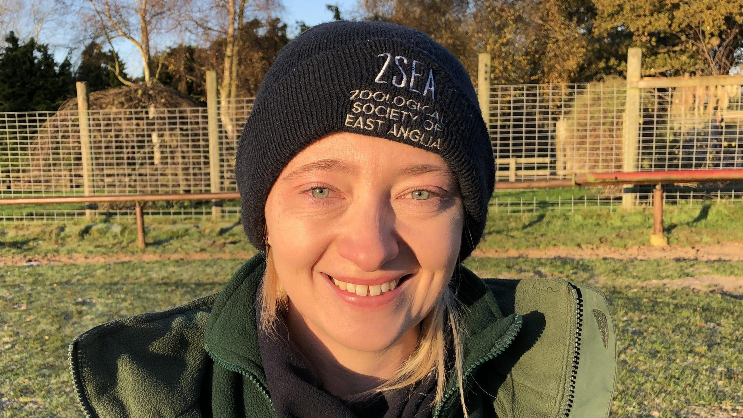 A woman smiles at the camera. She is wearing a black beanie hat with a black snood around her neck and she is in a green coat and green jumper. Large fences can be seen behind her.