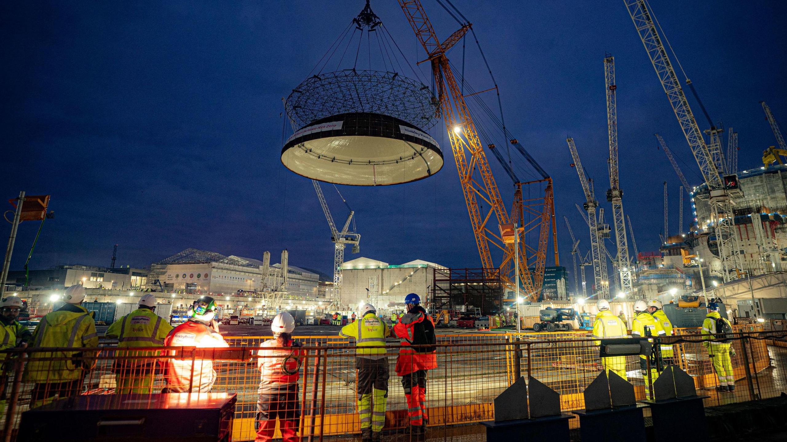 A huge dome is lifted high against the night sky by a giant crane, the biggest in the world. Building workers look on over a guardrail in the foreground, and there are dozens of cranes rising above the site.