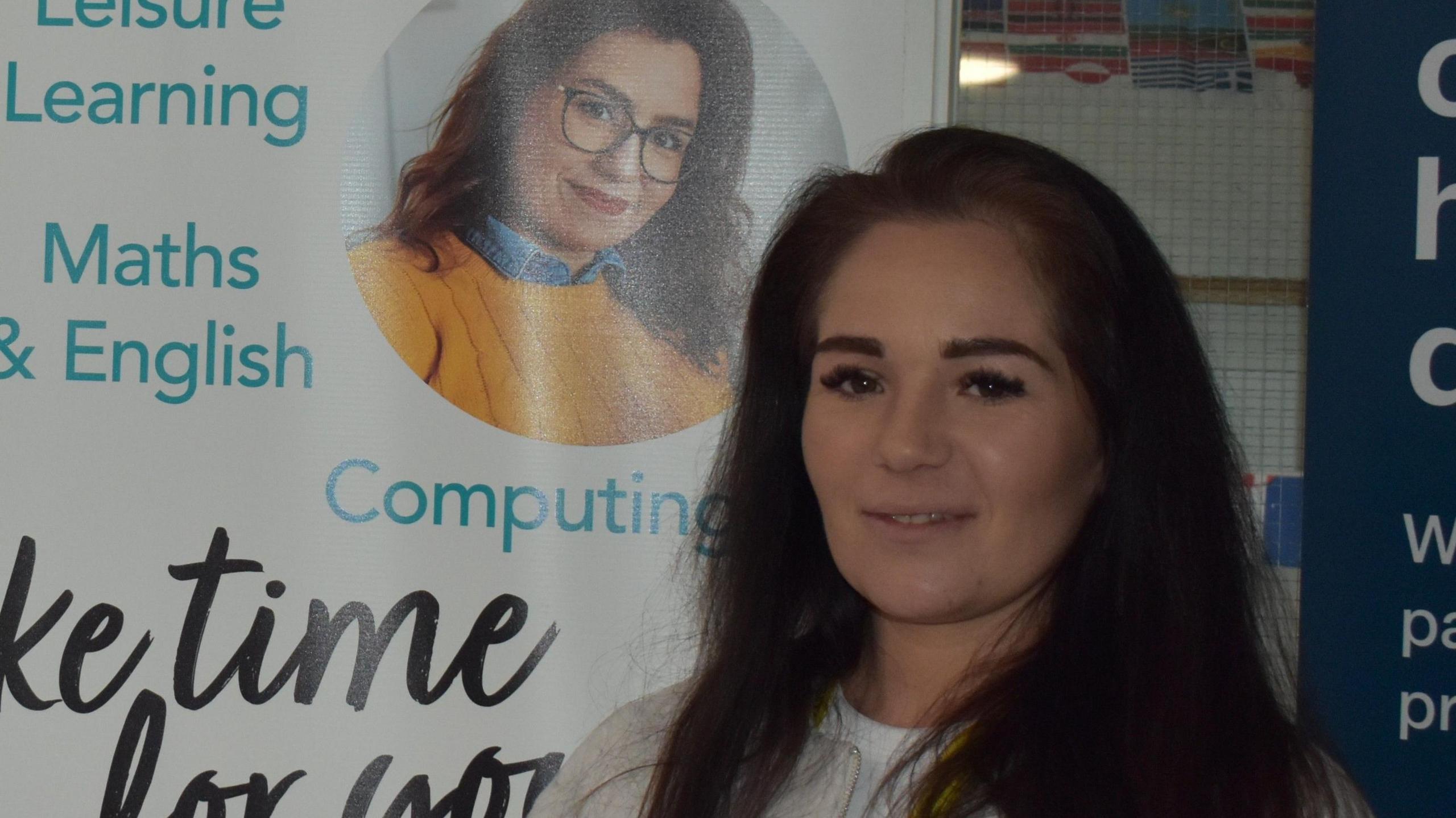 Ms Porter smiles at the camera. She has long dark hair and wears a grey jumper. She stands next to a promotional board inside a college.