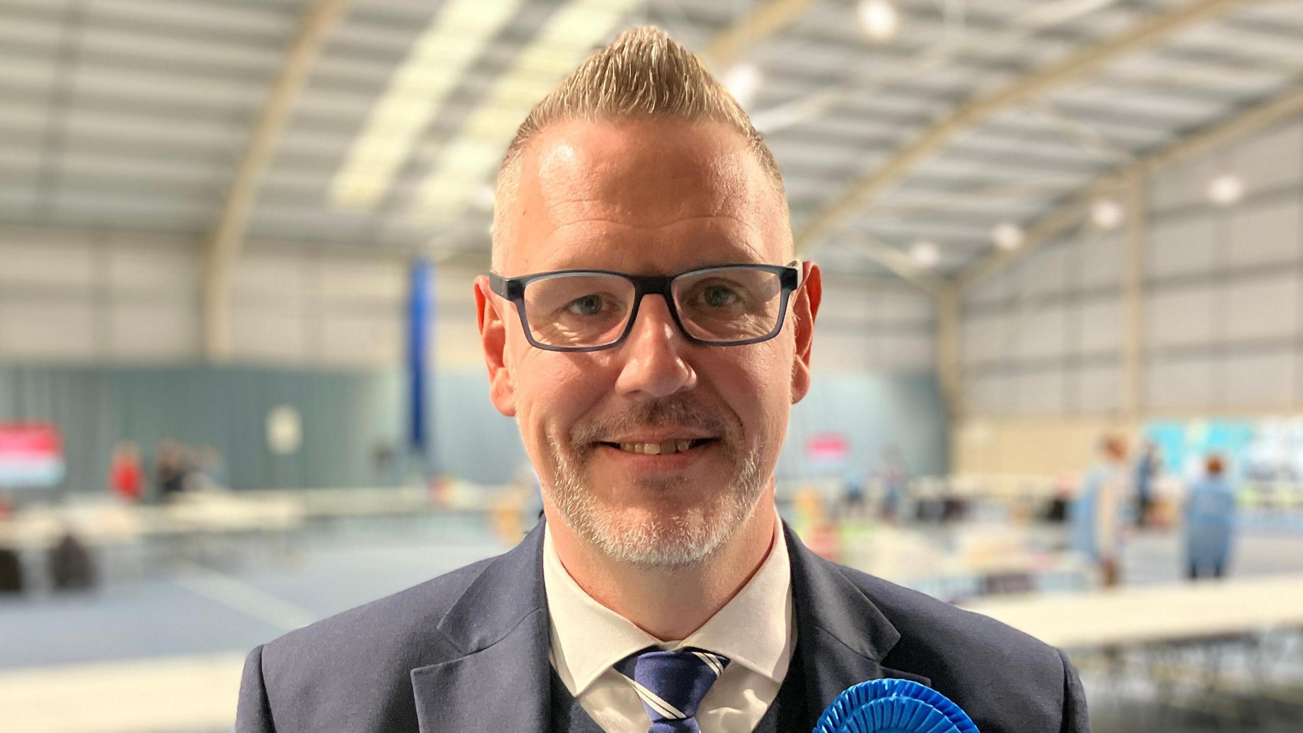A man with short, grey spikey hair is pictured in a sports hall wearing glasses and a navy blue suit. 