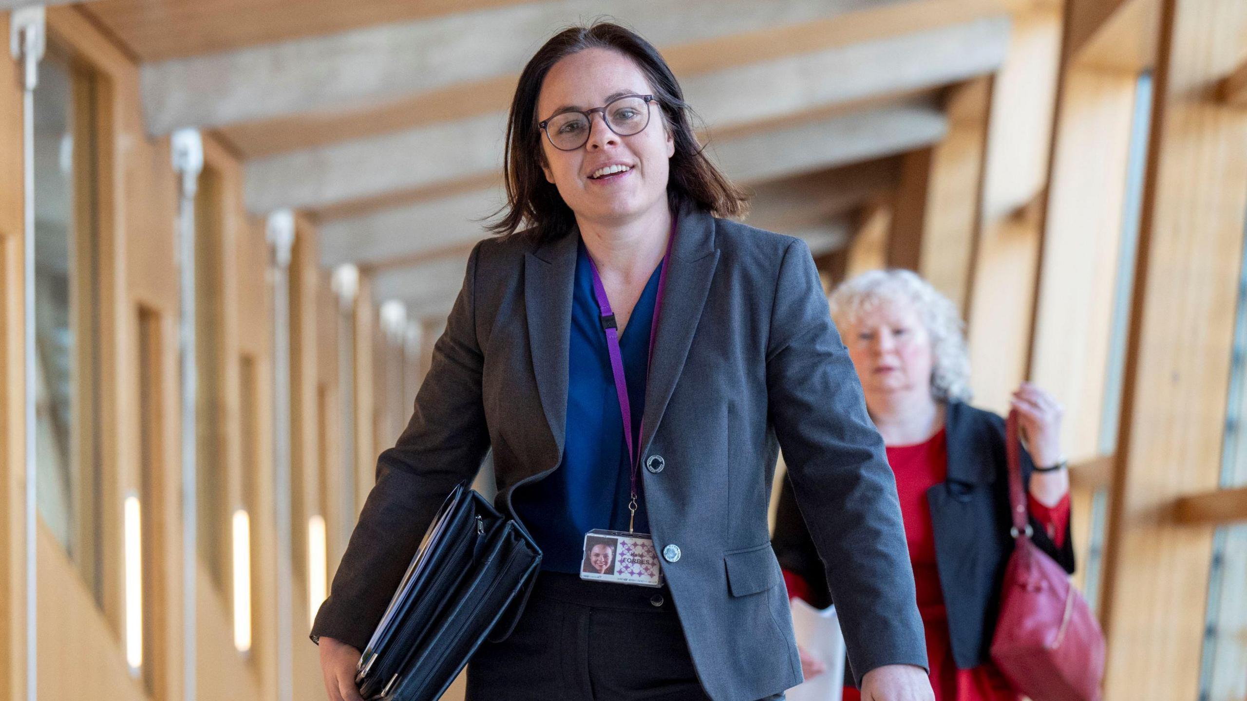 Kate Forbes walking in a corridor - she in a grey jacket, with a blue top, a lanyard round her neck