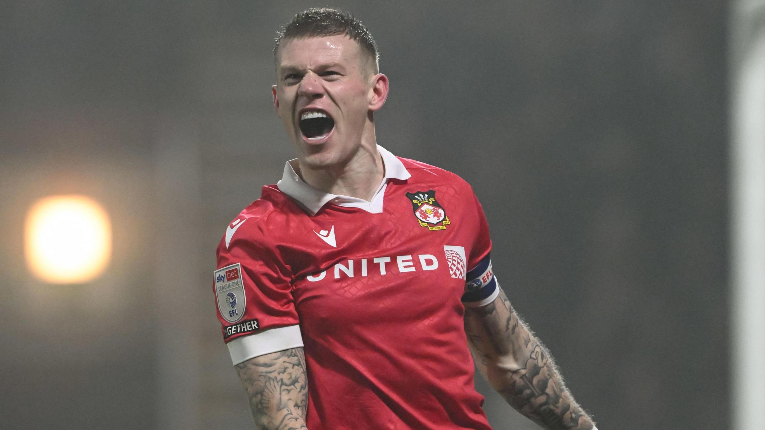 Footballer James mcClean pictured celebrating a goal. He is wearing the red strip of Wrexham FC, his arms are heavily tattooed