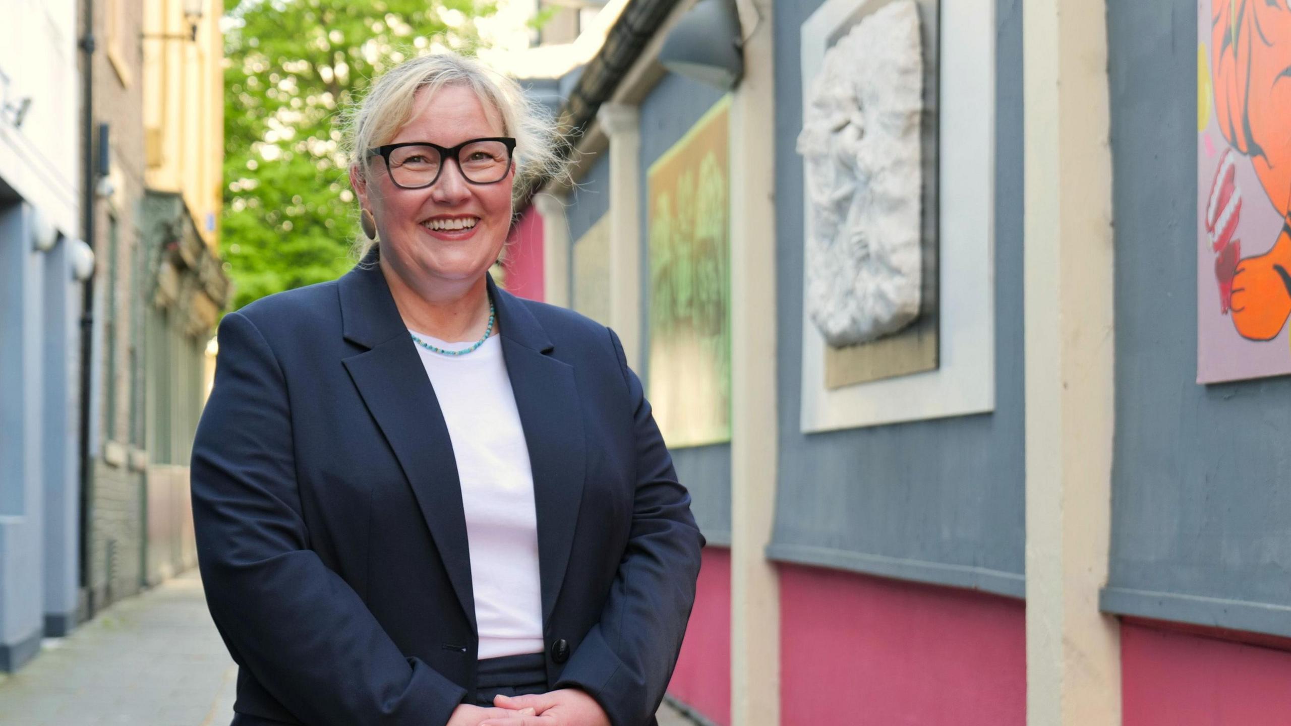 Newcastle City Council leader Karen Kilgour stands next to an art display outside smiling. She is wearing glasses and has blonde hair.