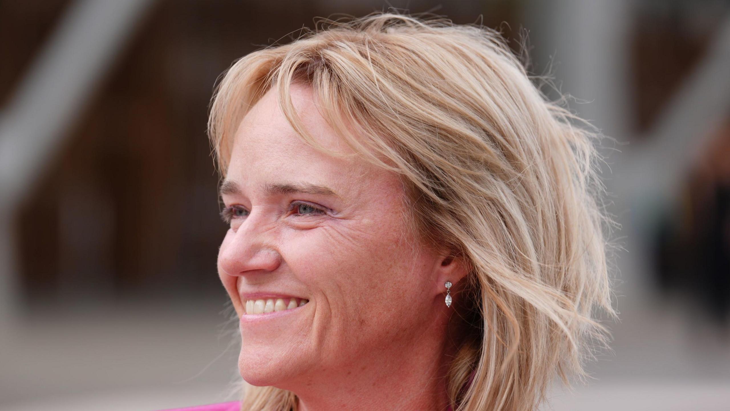 Sandie Peggie - a woman with blonde hair to her shoulders and a pink jacket smiles while standing outside
