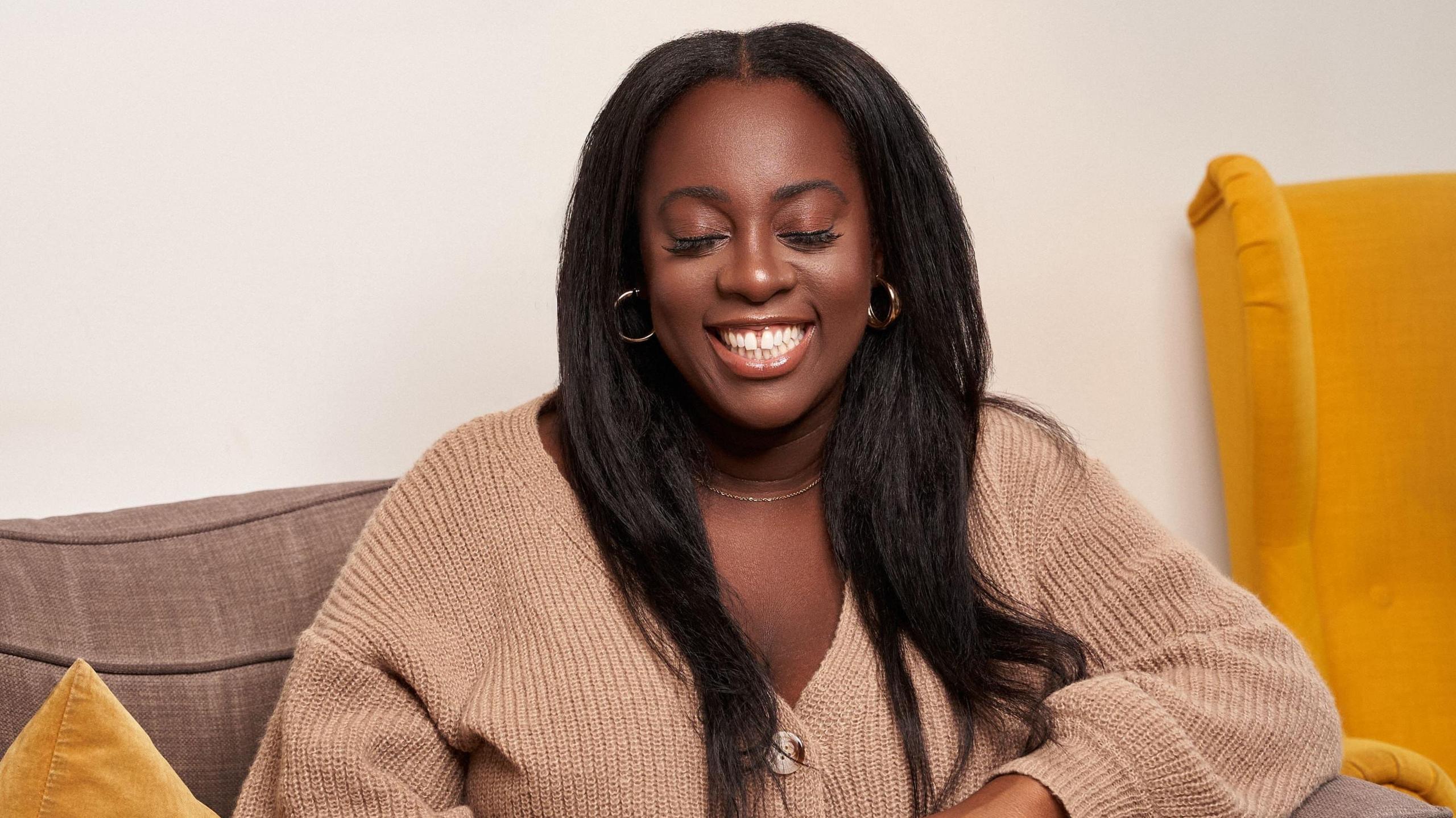 Jorden Berkeley pictured sitting on a sofa. Jorden has long black hair and wears a light brown cardigan. She smiles, closing her eyes. In the background there is a yellow chair.  