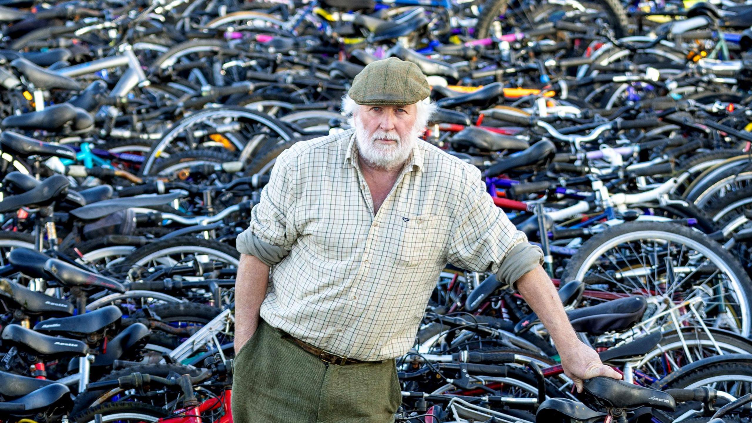 Nigel Carter leans against one bike as he stands amongst thousand while looking at the camera.
 He has white hair and a beard, wearing a checked shirt and green trousers. 