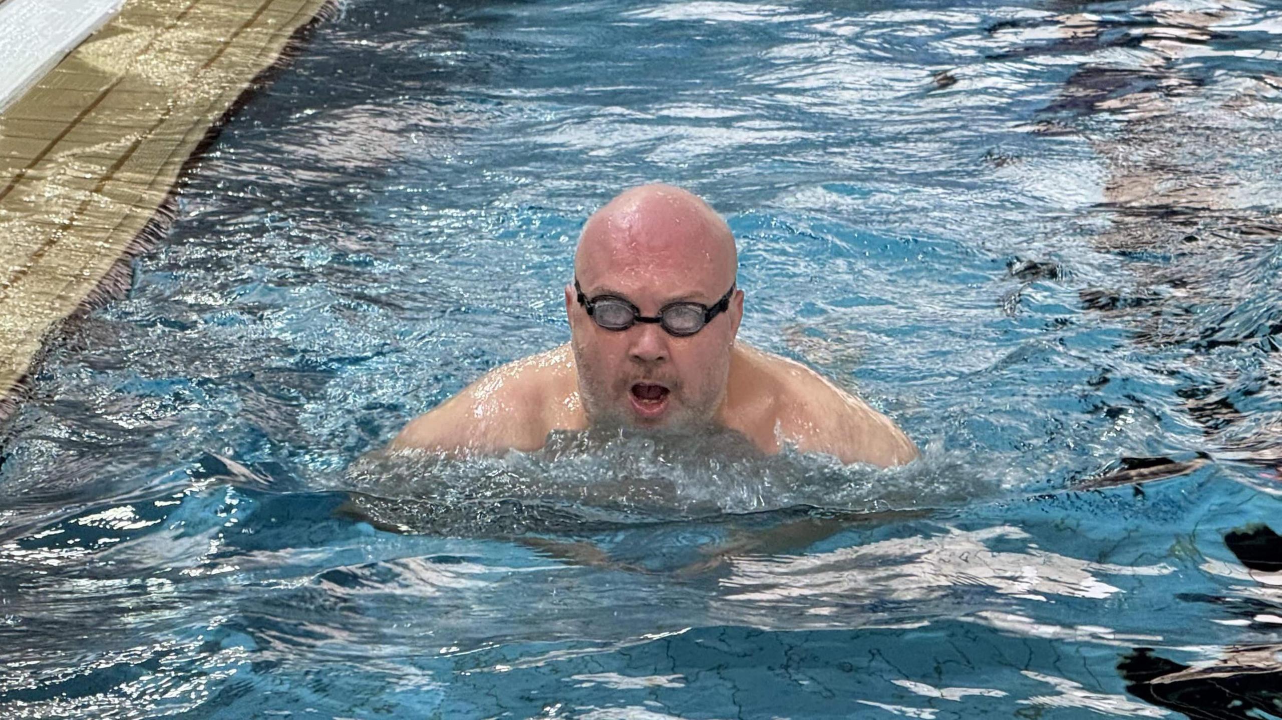 Radio presenter Richard Stead swimming in a pool. He is wearing black goggles and is staring towards the camera. He has a bald head and a grey beard.