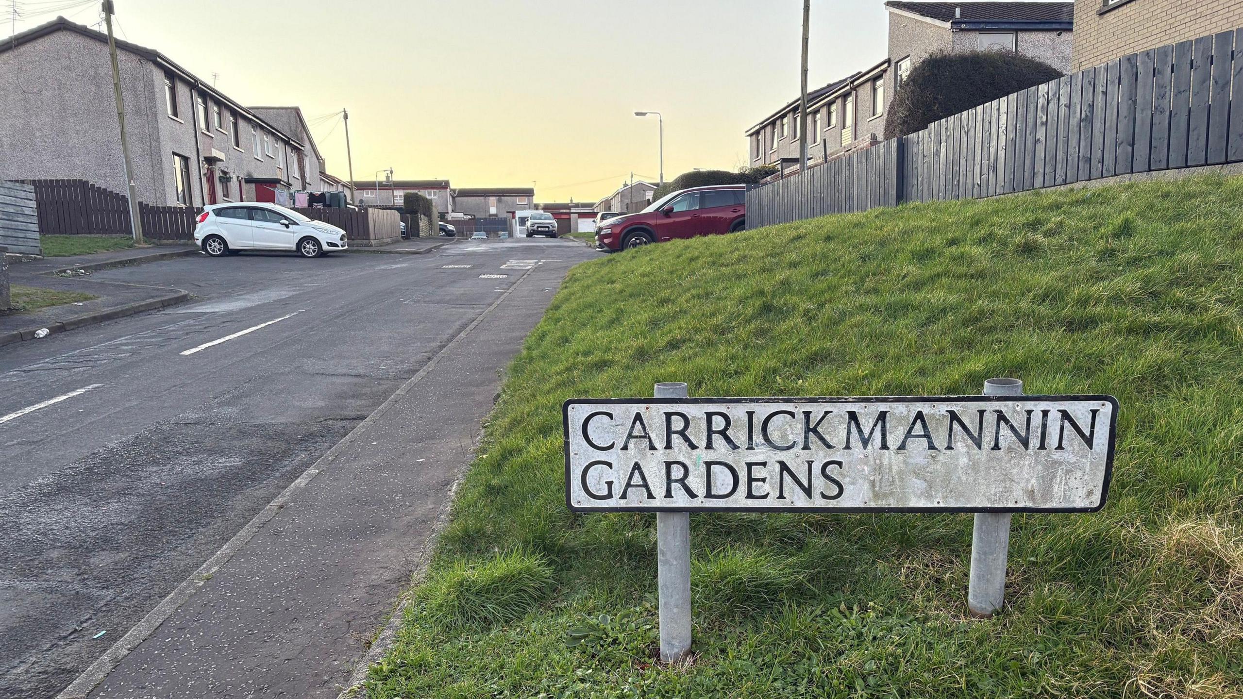 A sign reading ' Carrickmannin Gardens' on a grass verge. Houses and cars can be seen in the background.