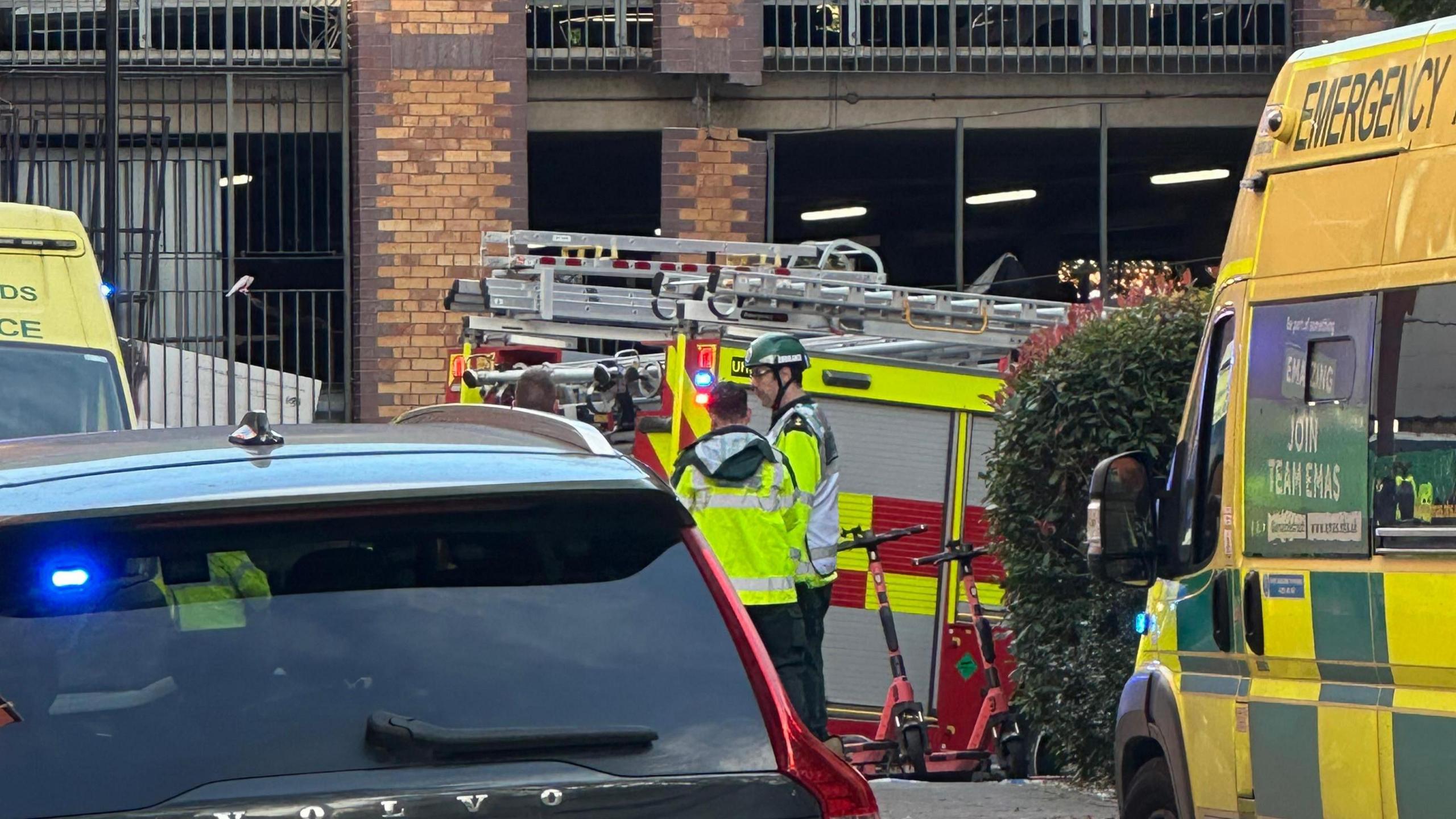 Two ambulances, a first responder car and a fire engine in front of a brick-built multi-storey car park. Two emergency services personnel are visible in yellow hi-vis.