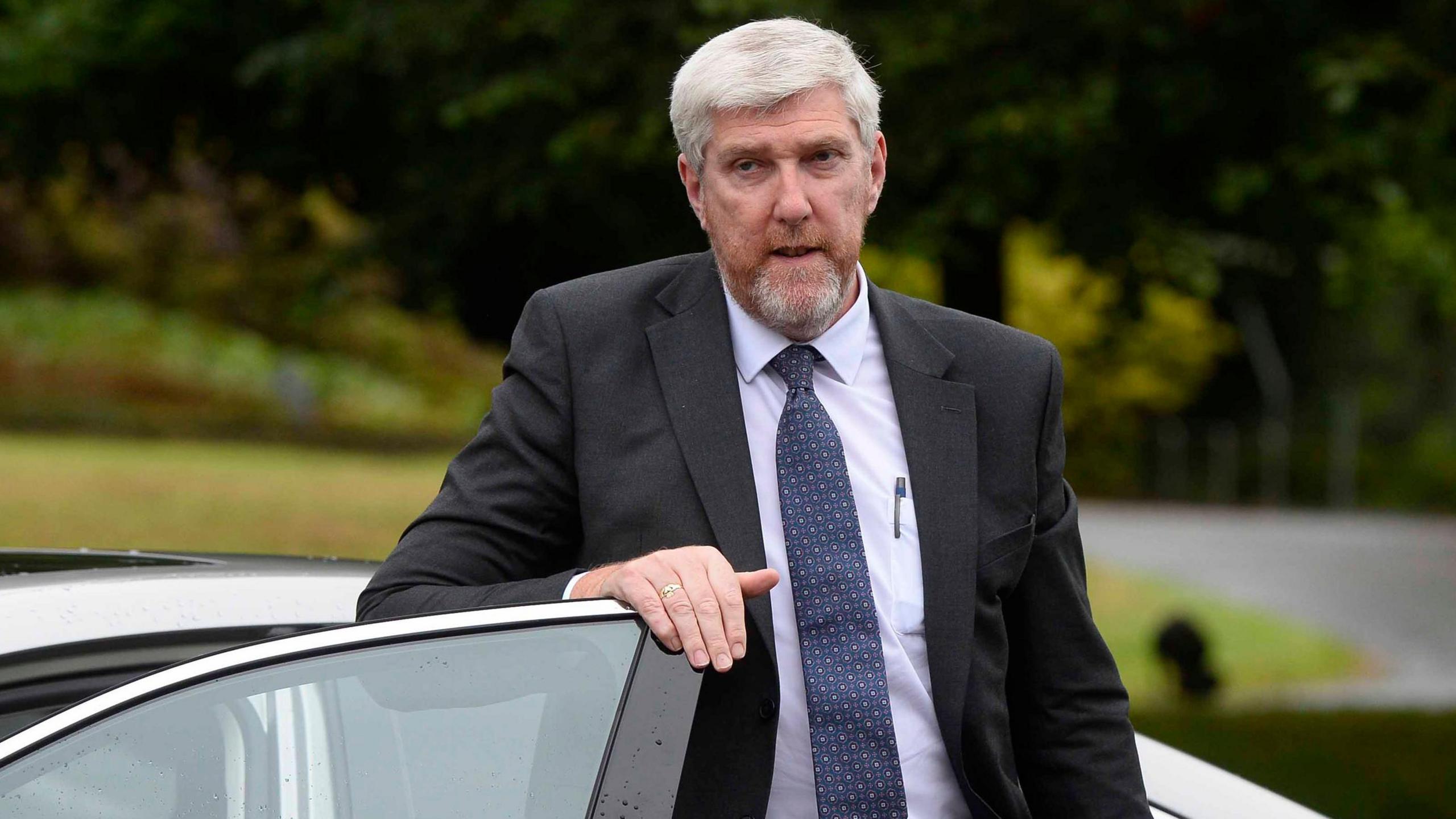 John O'Dowd is getting out of a silver car with leafy background of trees, grass and shrubs. 
He has silver grey hair and a gingery, silver beard.
He is wearing a navy blue tie with pink spots on it.
He has a white shirt with a pen in his pocket. 