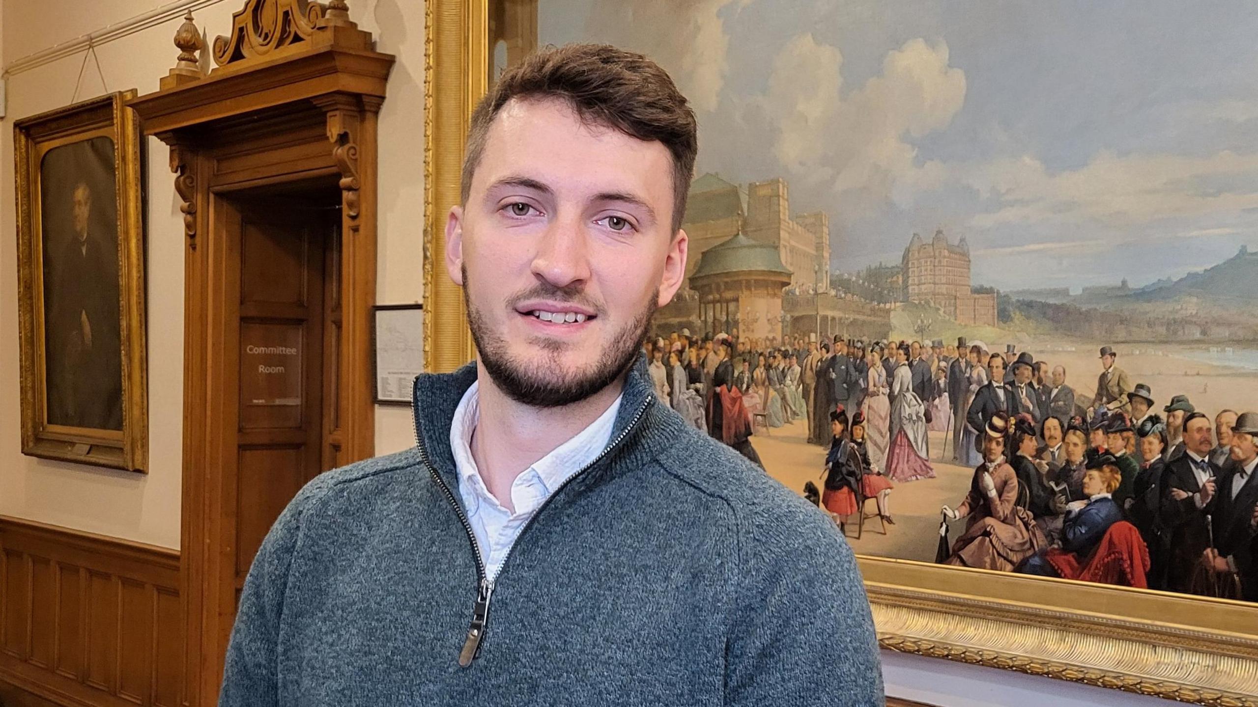 A man in a grey quarter zip fleece stands in front of a large painting in a council building.