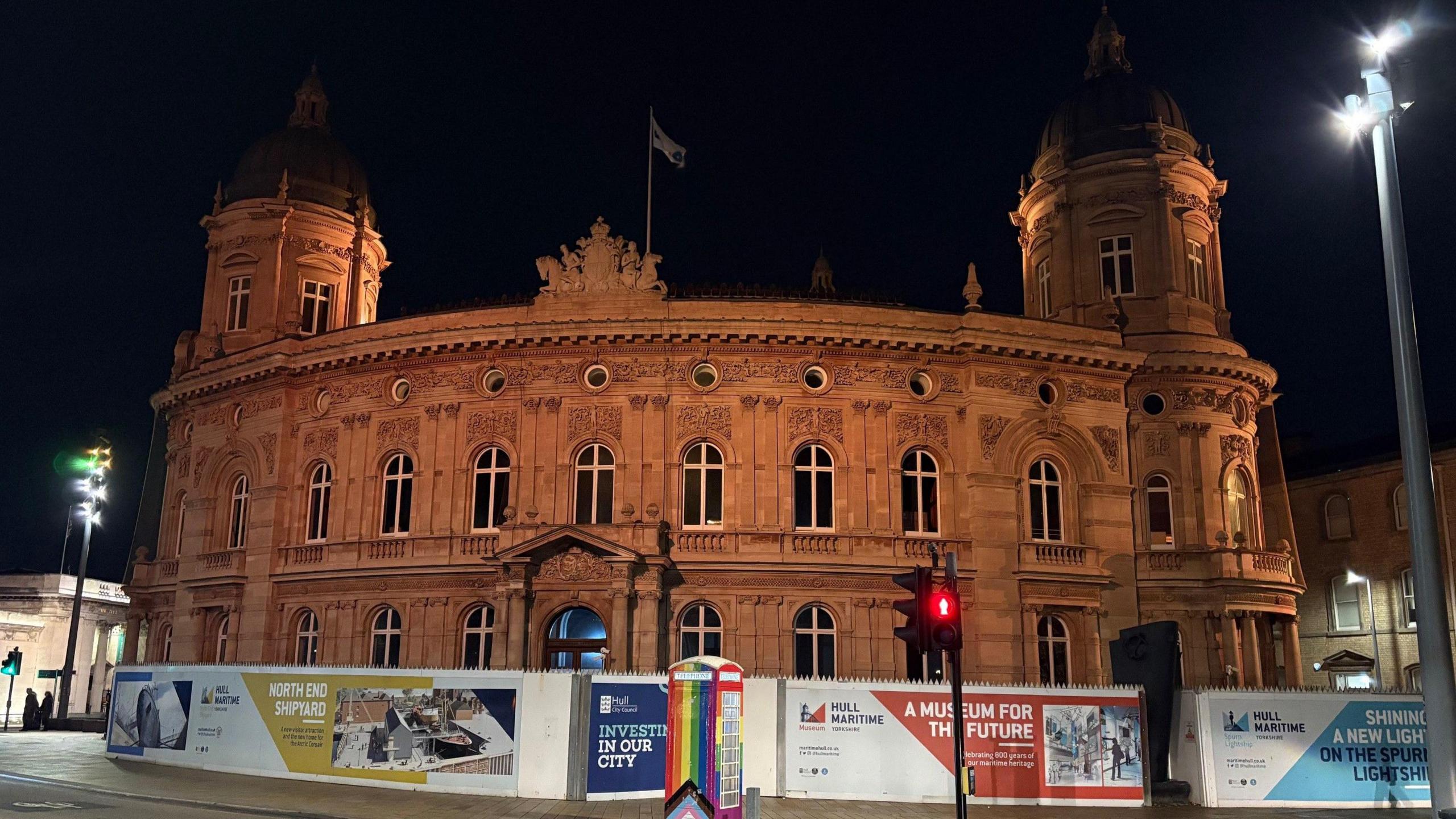 A large building, made from sandstone, glows in the light from street lamps. In front of it, are hoardings promoting Hull City Council's significant restoration project.
