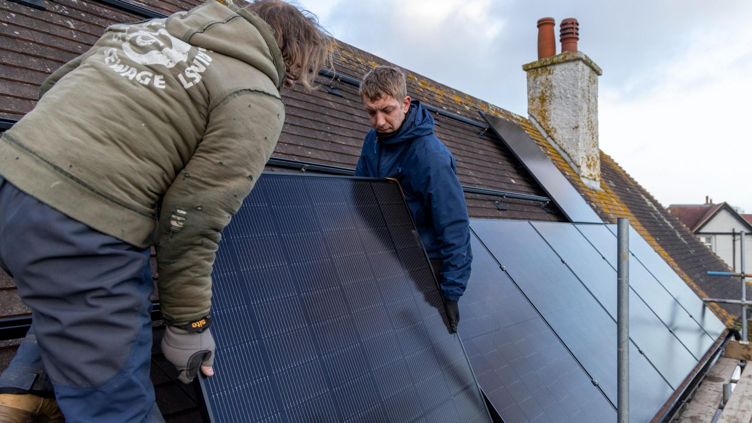 Two men install a solar panel on a brown tiled roof. Five panels are already laid behind. The man on the left has his back to the camera, he is wearing a green jumper and blue trousers, the man on the right facing the camera wears a blue jumper