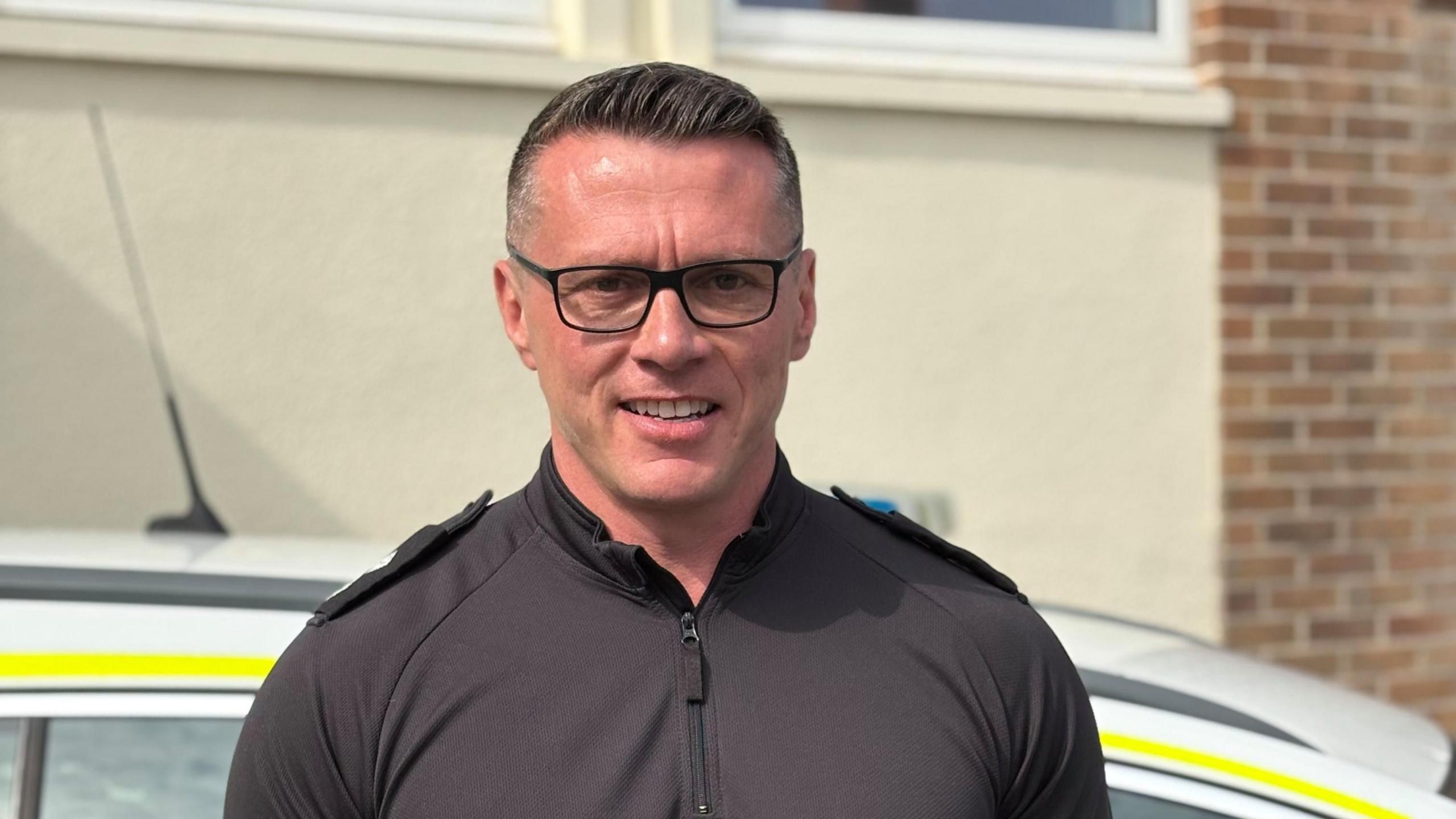 Inspector Terry Murphy smiles at the camera while standing in front of a police car. He is wearing black police uniform and black rimmed glasses with short hair that is gelled.