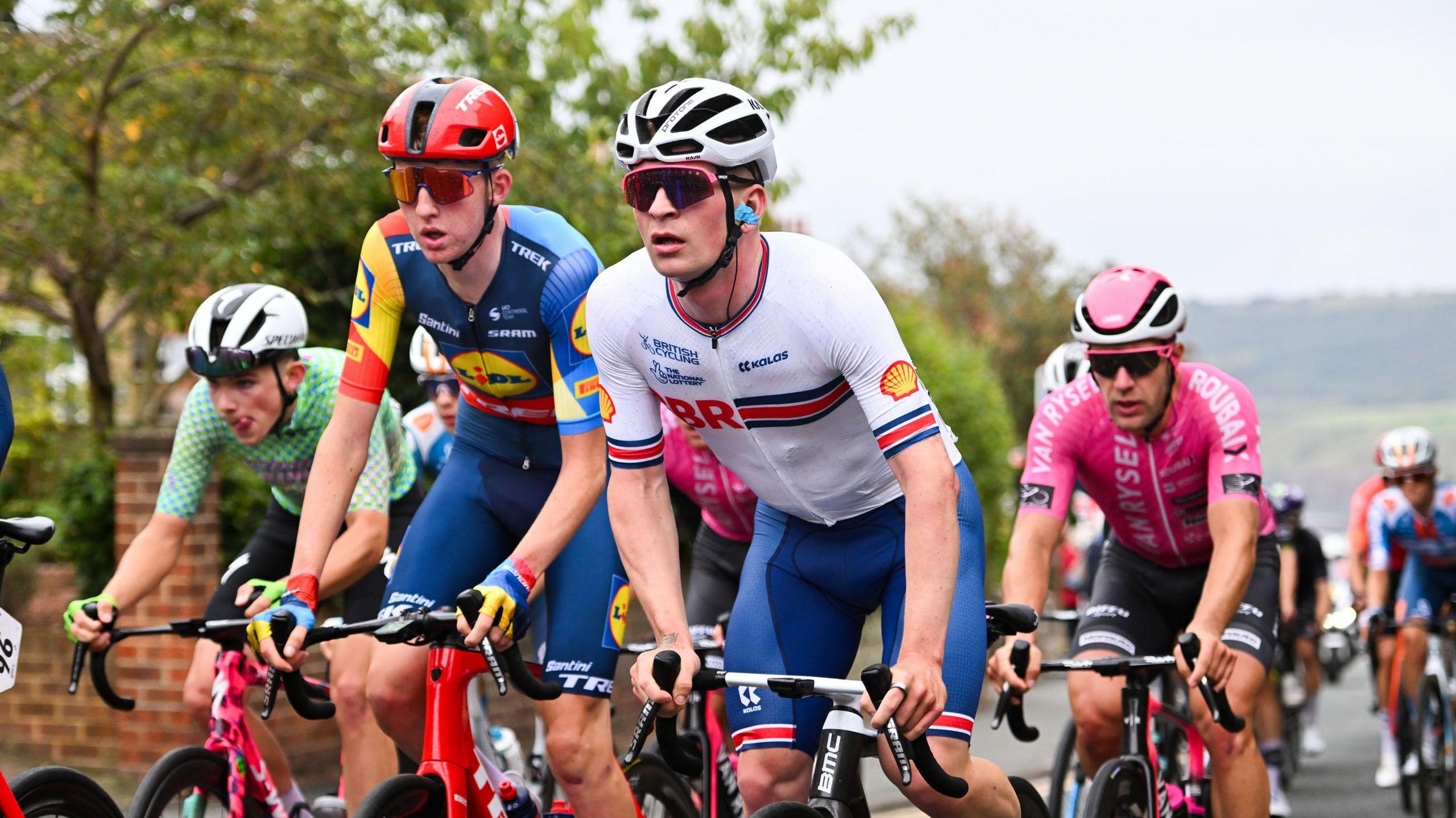 Four professional cyclists racing against each other on a road. Other cyclists can be seen in the background.