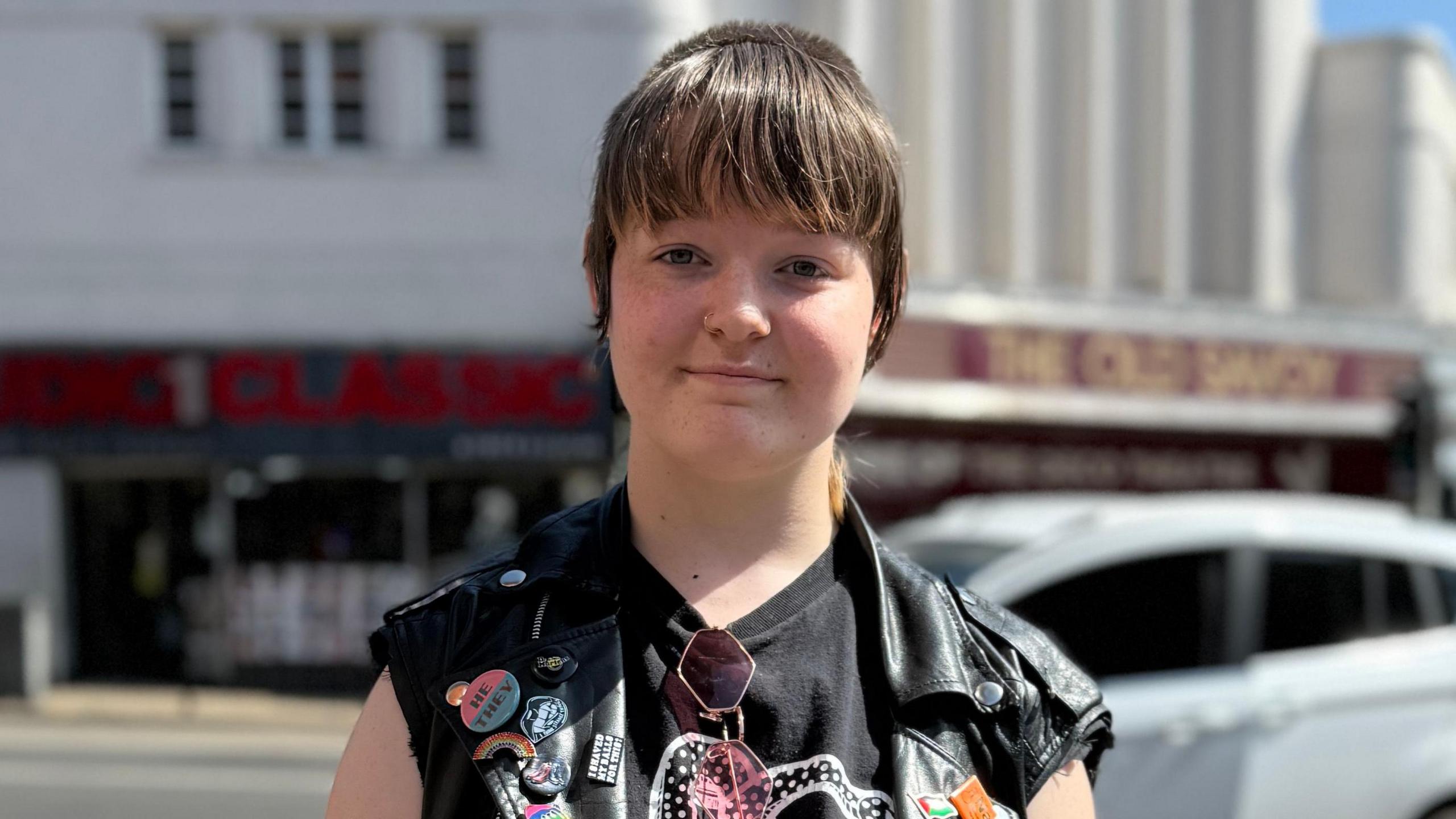 Bowie is standing outside. They have a fringe and are wearing a black T-shirt and black sleeveless jacket. They are looking directly at the camera and smiling.