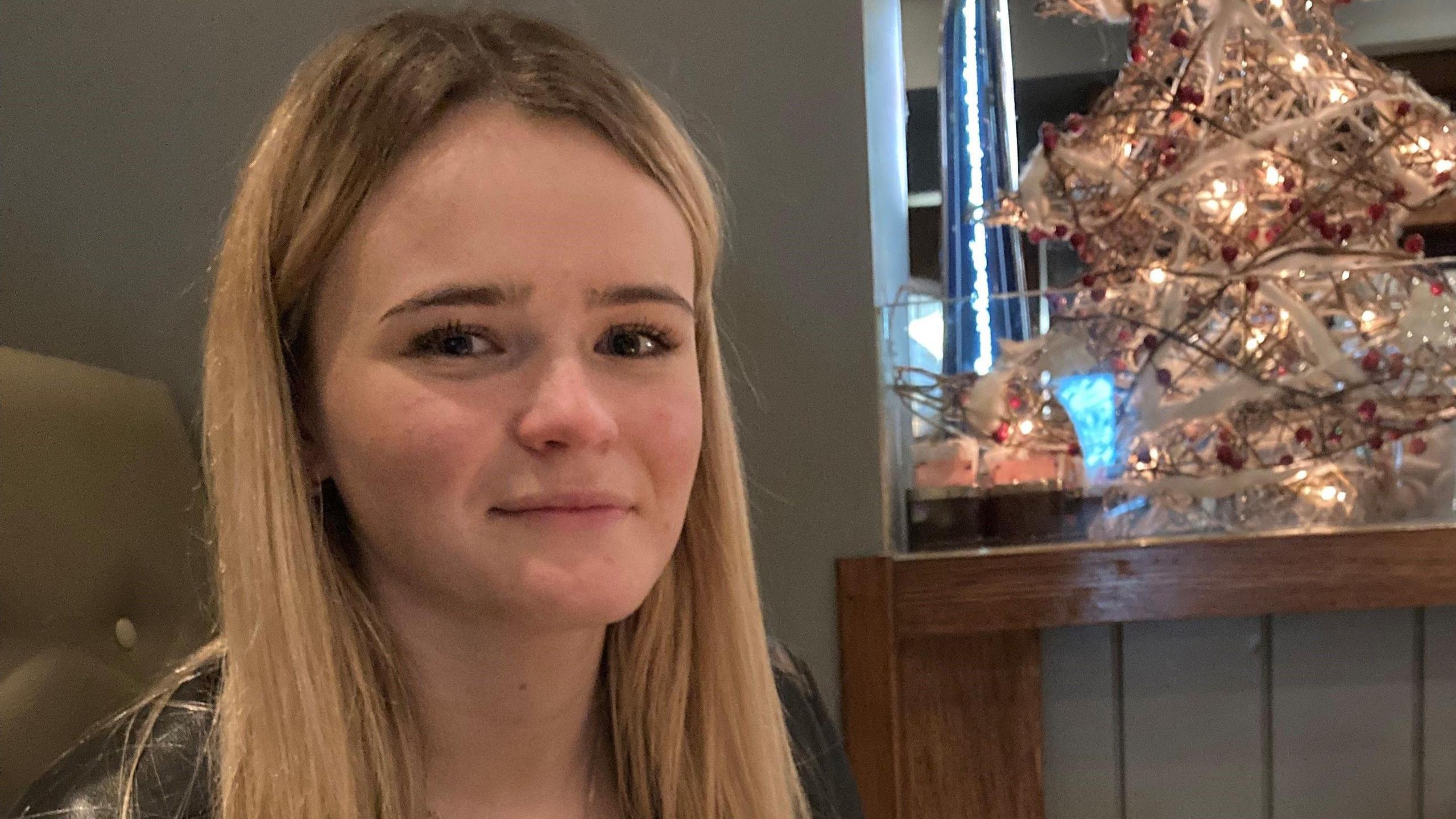 Amelia Butlin with blonde hair, gazing at the camera with a small faux Christmas tree in the background. 