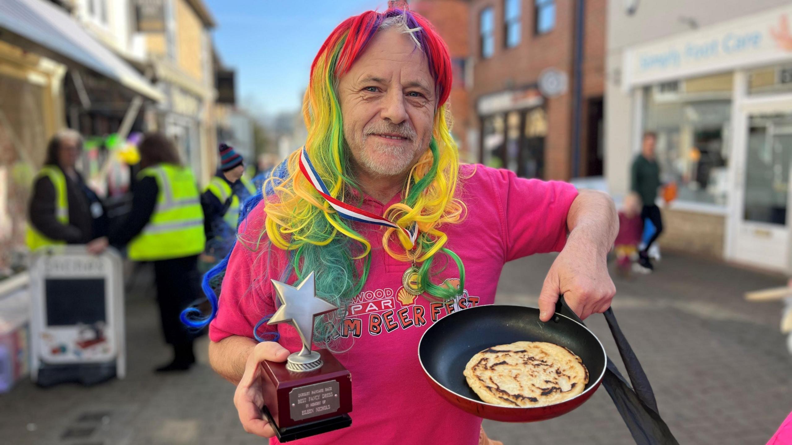 A man in a pink t-shirt and a colourful wig holding a trophy and a pancake in a pan after the annual fancy dress pancake race in Dursley