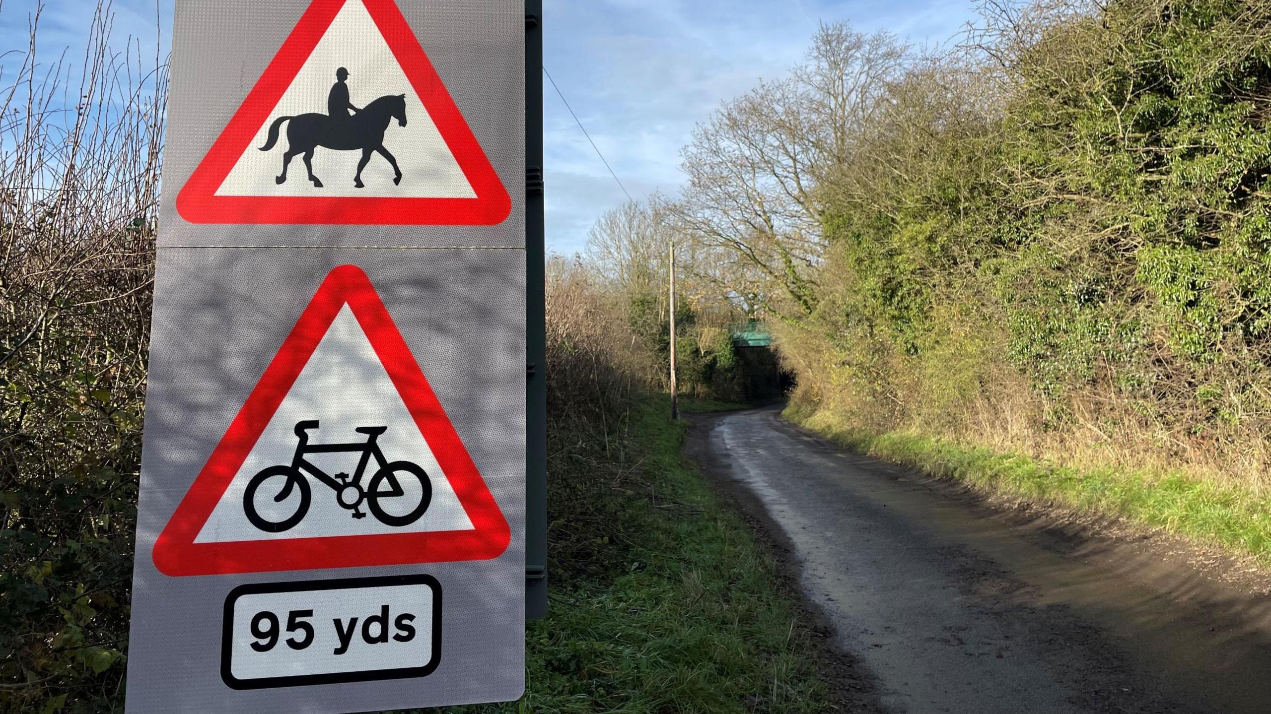 A photo of Batley Lane, in Pleasley. A thin single-track road can be seen on the right. On the left are two triangle road signs of a person riding a horse, and another of a bicycle