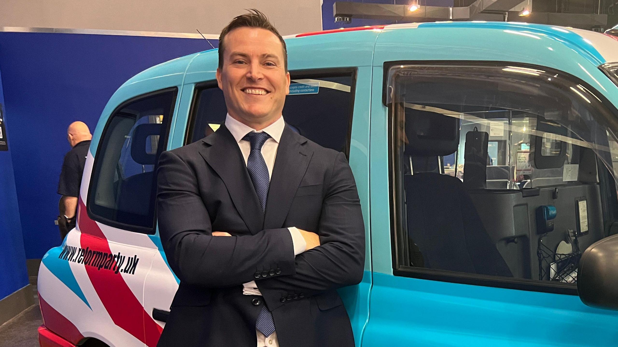 James McMurdock is standing in front of a Hackney cab carriage, which is painted in the colours of Reform UK (blue, white and red). He is wearing a navy blue suite and a blue tie.