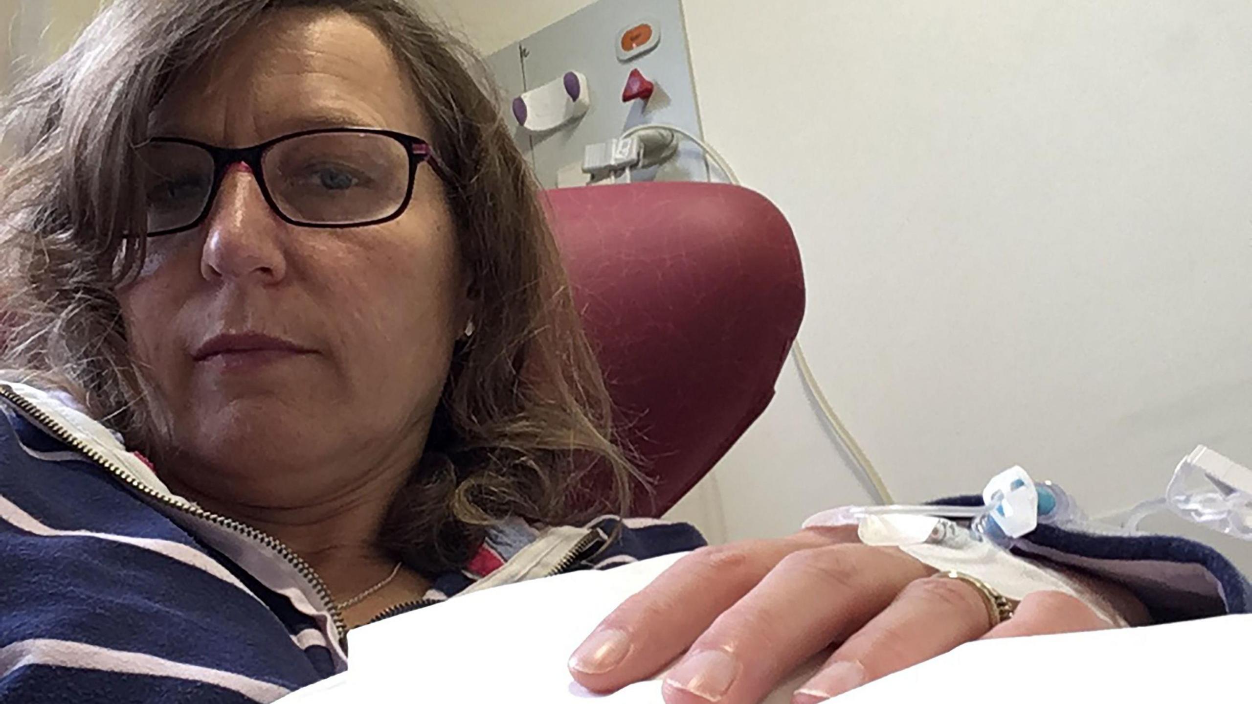 Sam looks at the camera, wearing glasses and a stripy top while getting treatment - there is a hospital line into her hand.