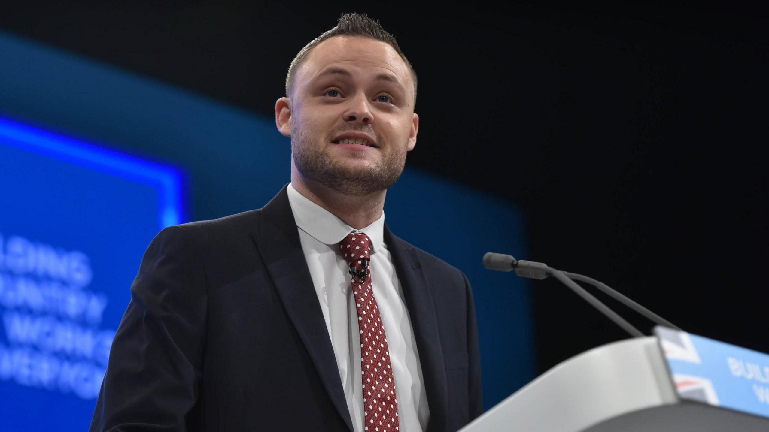 The Conservative leader of Nottinghamshire County Council Ben Bradley speaking at a podium at a conservative conference wearing a suit with a black jacket and a red polka dot tie