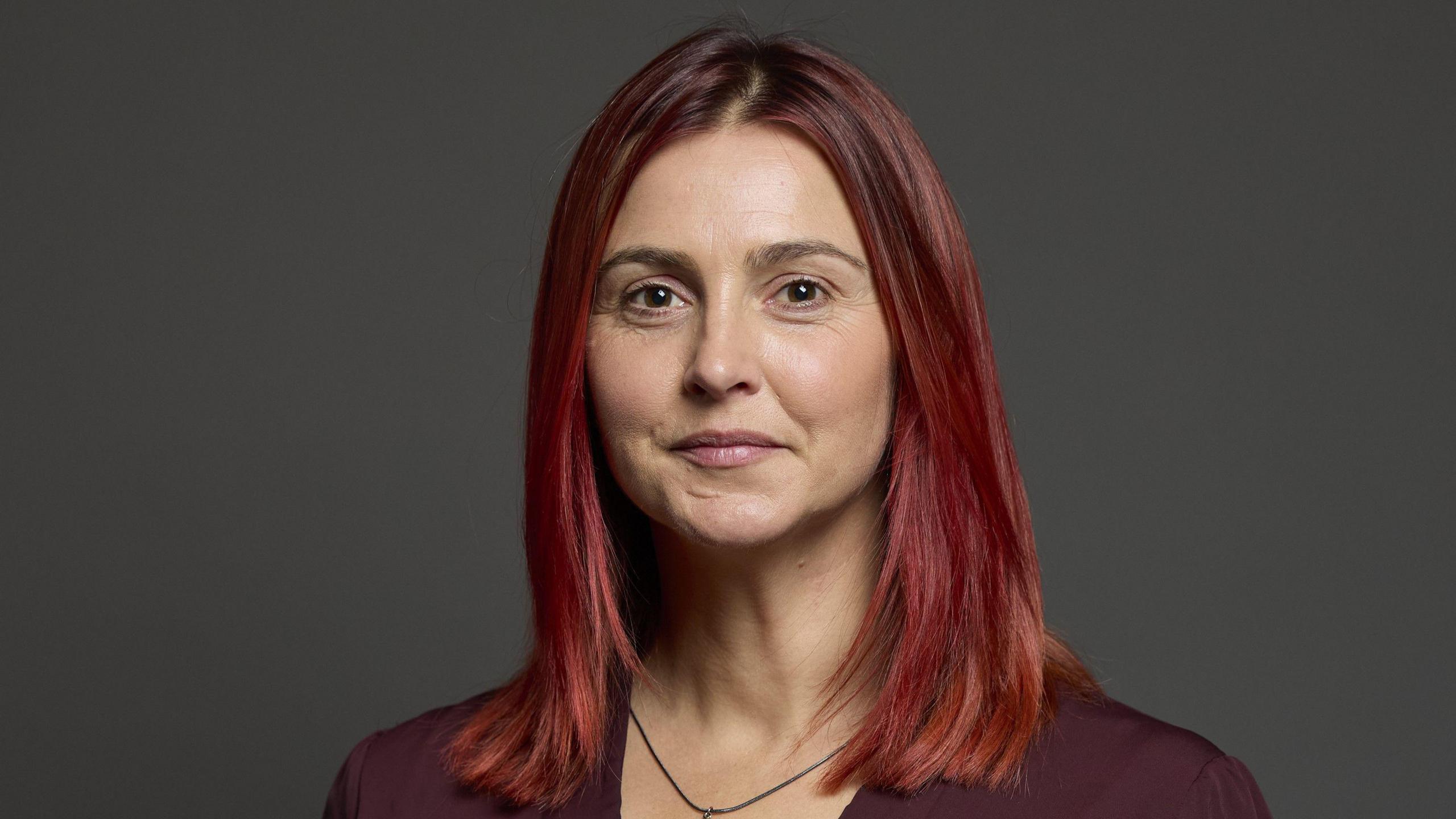 A woman with bright red shoulder-length hair, wearing a purple blouse and necklace.