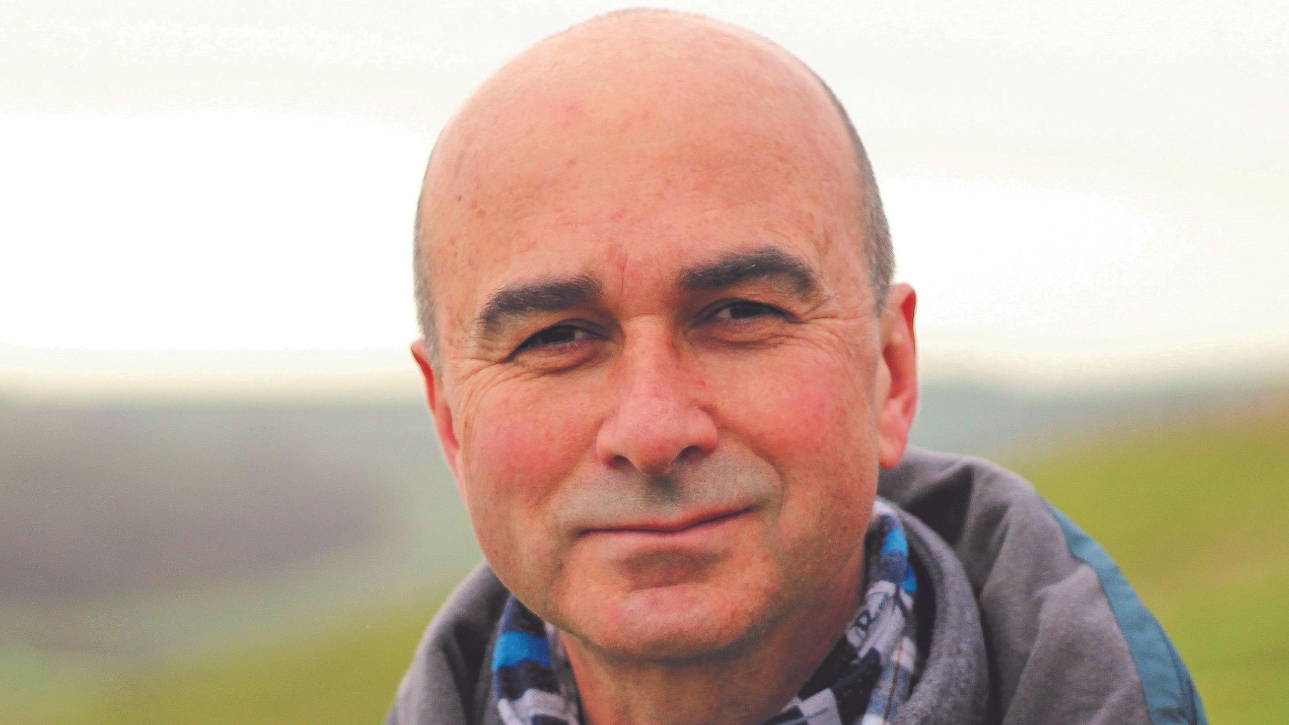 A man looking straight at the camera with a slight smile. He is standing in front of green fields and is wearing a grey and teal coat and a black, grey and blue checked shirt.