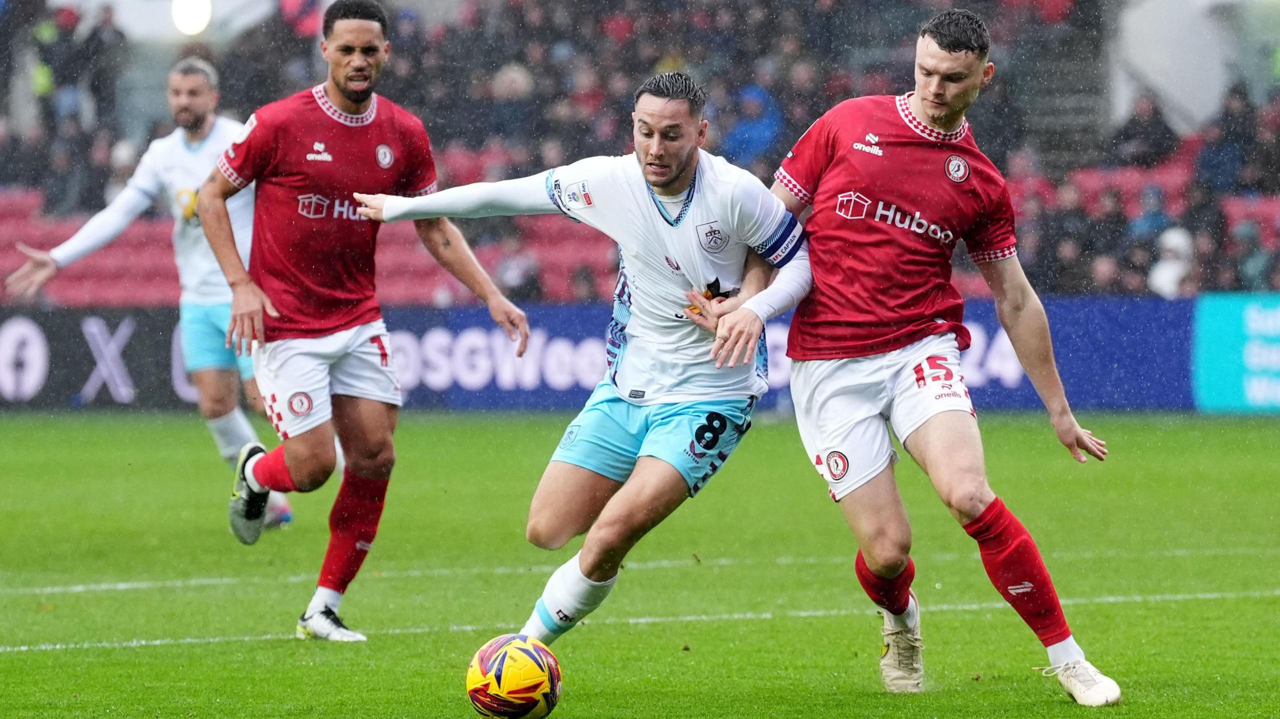 Players from Bristol City and Burnley contest the ball at Ashton Gate. Heavy rain can be seen falling around them. Bristol City are in their traditional red shirts and white shorts while Burnley are in their away strip of light blue shorts and white shirts