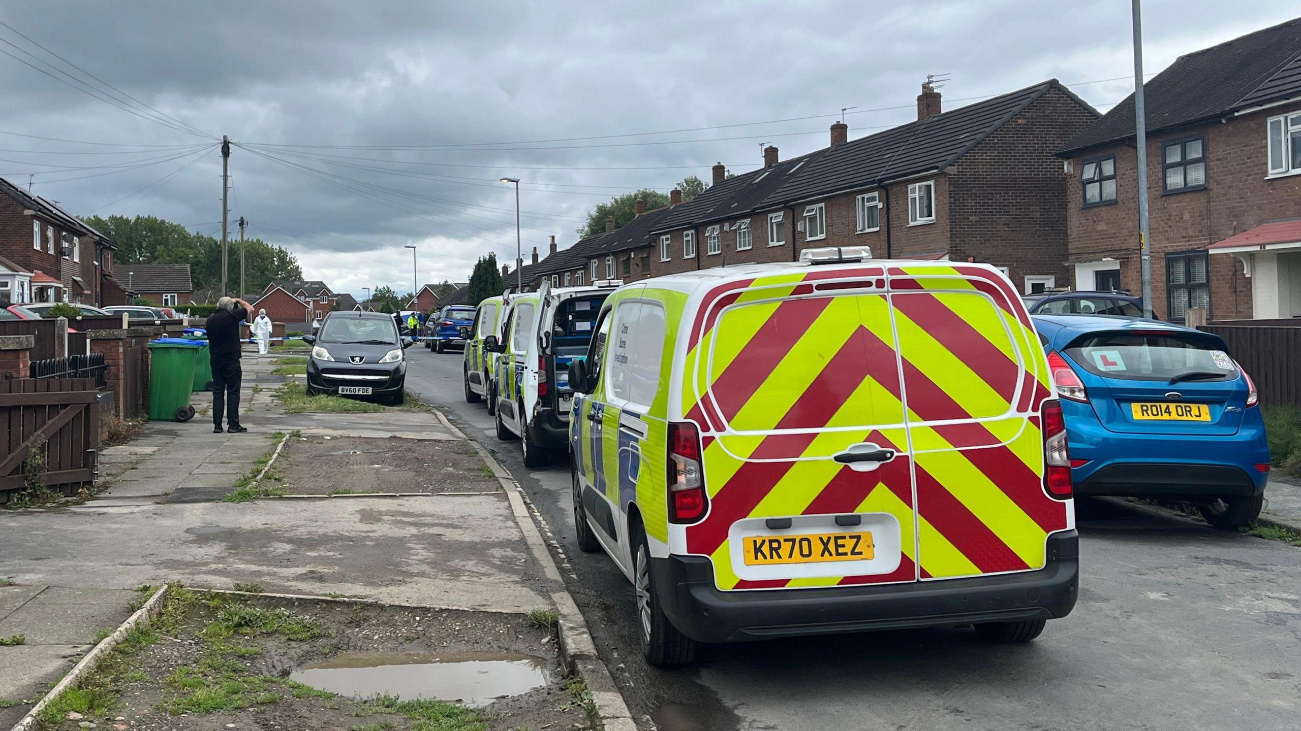 Three police vans are parked in a row on a residential street. A man wearing all black stands on the footpath taking a photo with a camera. In the background, a forensic investigator wearing a full white suit and hairnet is stood behind police tape.