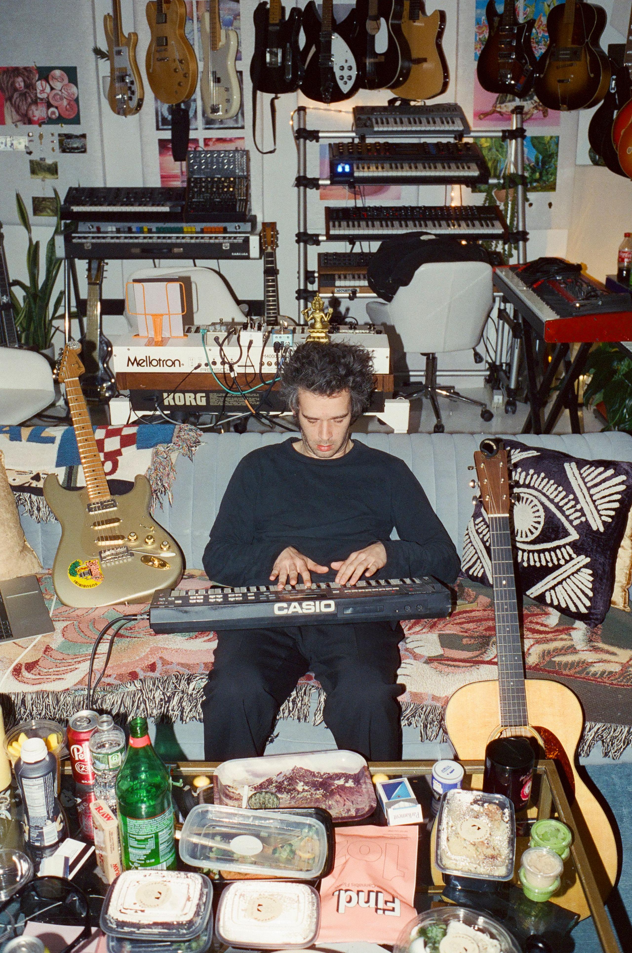 Matty Healy from The 1975 plays a keyboard while surrounded by clutter and musical instruments in what looks like a living room